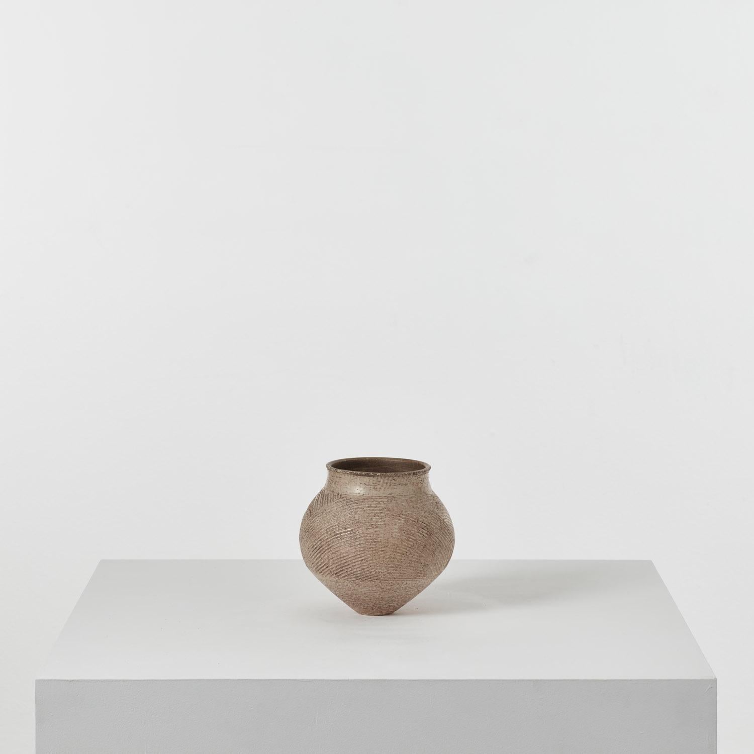 Soft striations of line subtly ridge across this Jason Wason pottery vase. Small at its base, the vase extends into a bulbous form, finishing with a circular top. This vase is a careful study of tactility, where the clay’s materiality and shifting