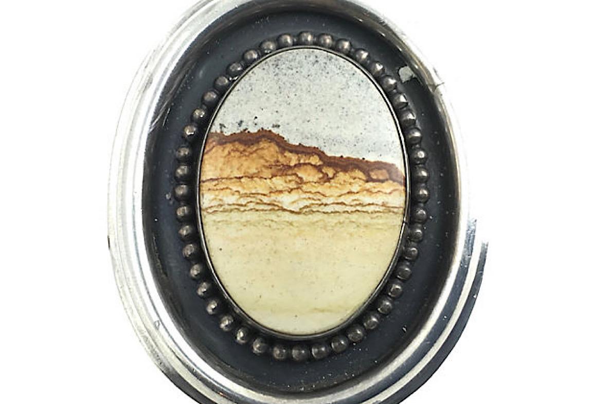 This Owyhee landscape brooch features a window scene of a desert sunrise with mountains in the distance on a piece of jasper mined from the Idaho-Oregon border area. The jasper is mounted in a simple oval sterling silver frame with a beaded border