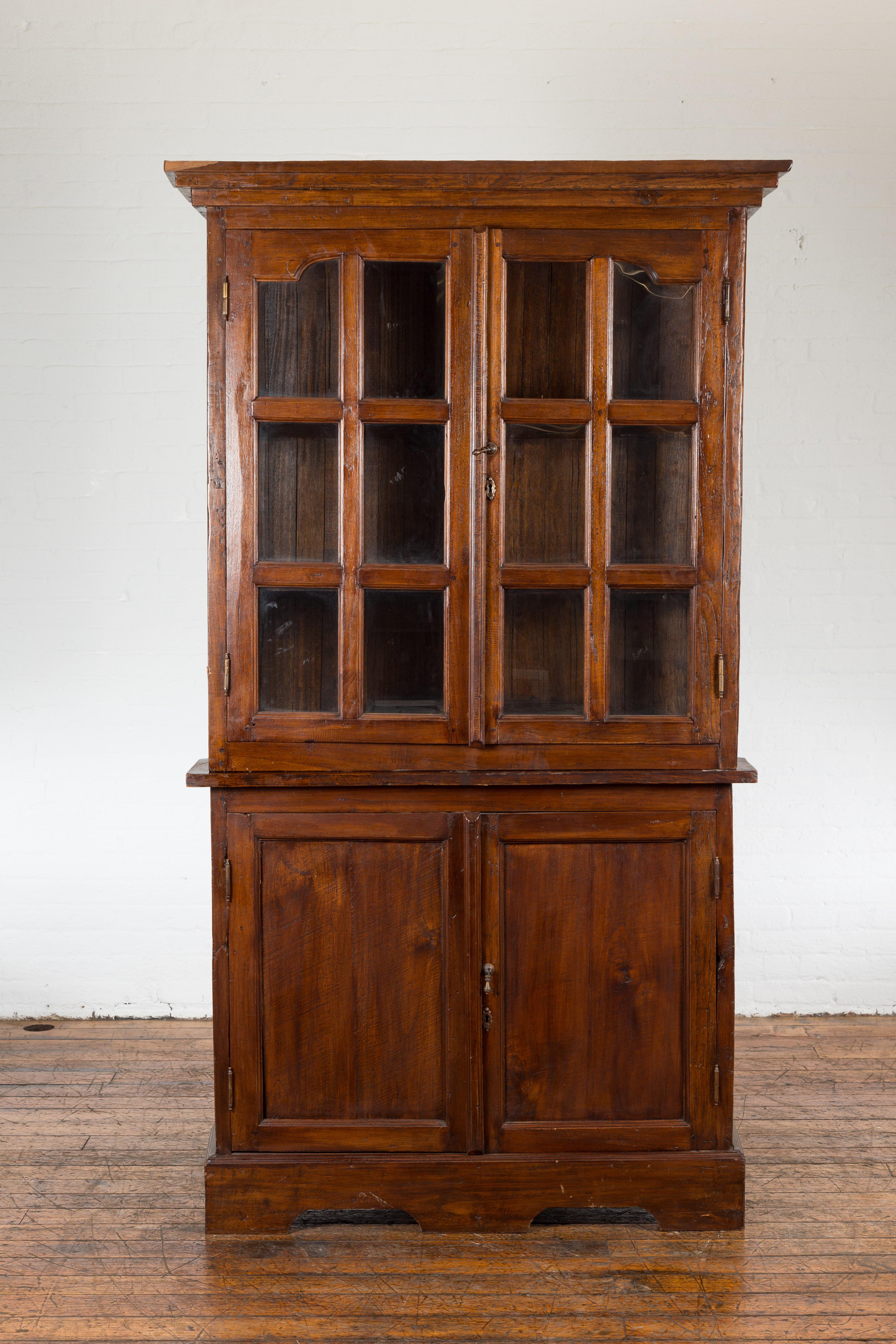 An antique Javanese Dutch Colonial period two-part display corner cabinet from the early 20th century with paneled glass doors in the upper section and wooden doors in the lower one. This antique Javanese Dutch Colonial corner cabinet from the early