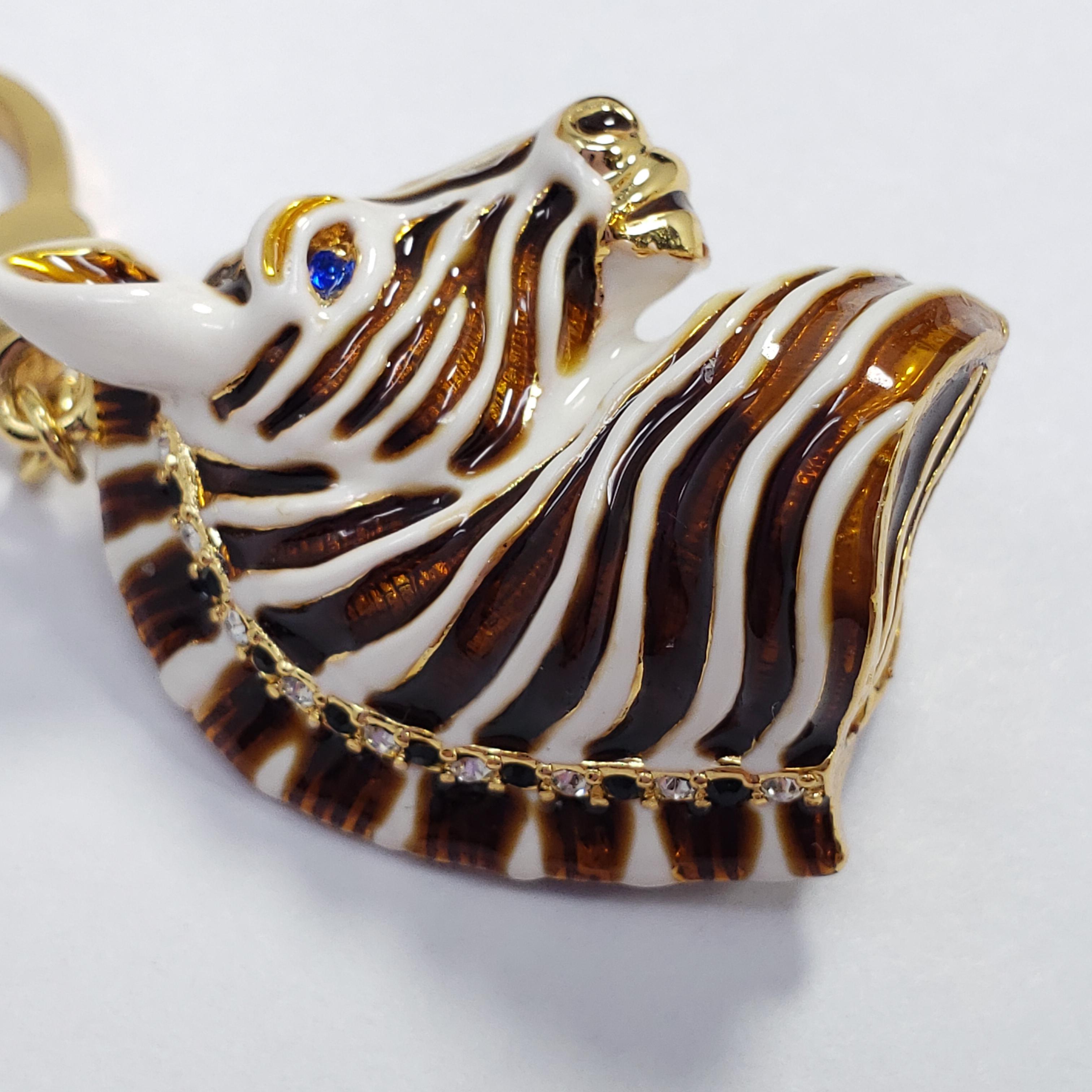 An enamel-painted white and brown zebra head is set on a gold tone metal and accented with clear and blue crystals. A whimsical charm, fitting as a pendant or any other creatively-styled accessory.

Originally for Jay Strongwater's line Jay, made