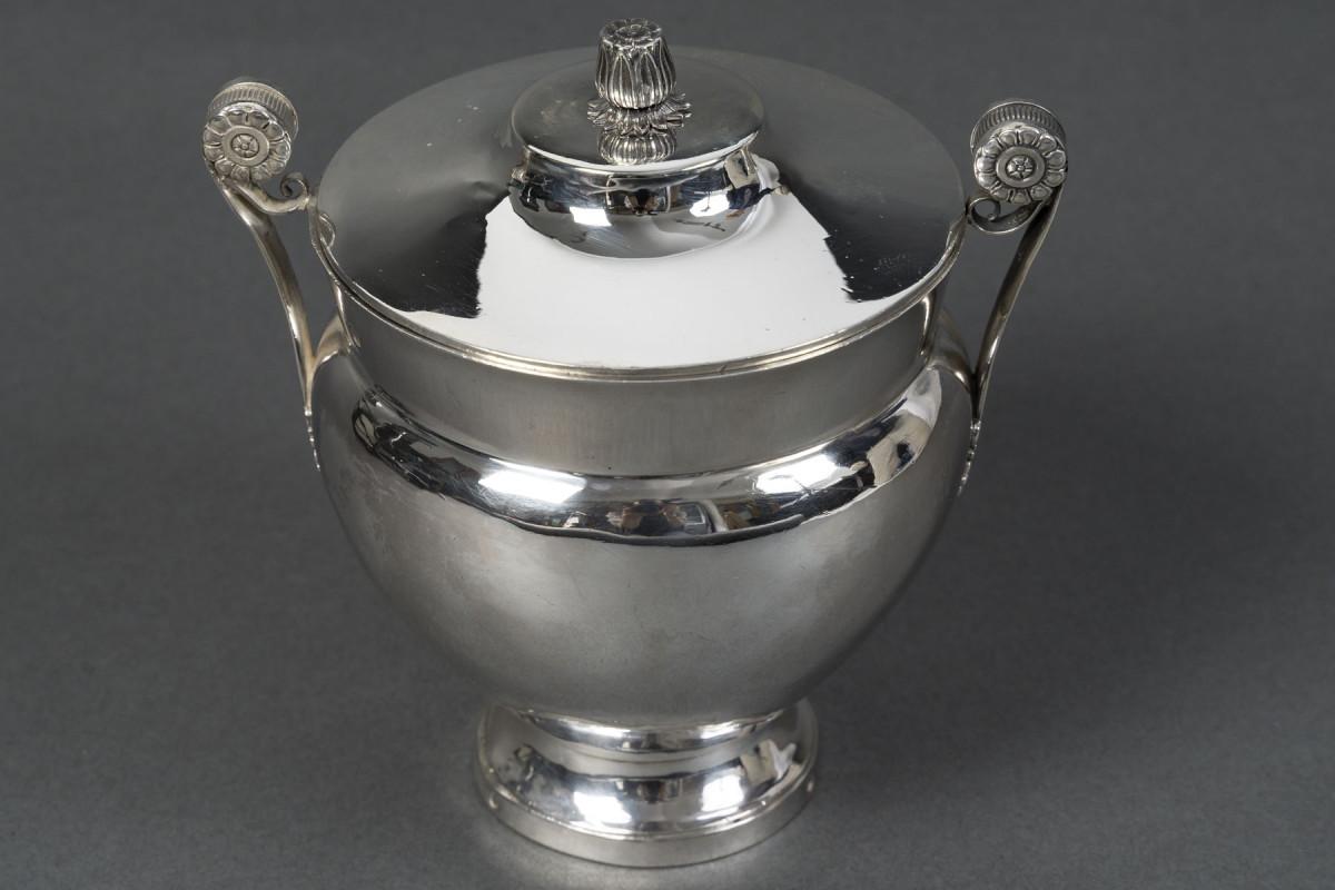 Elegant egg-shaped covered sugar bowl in solid silver, resting on a circular pedestal, with a domed lid adorned with a stylized seed handle. The side handles, finely chiseled with palmettes and rosettes on a matte background, reflect a decorative