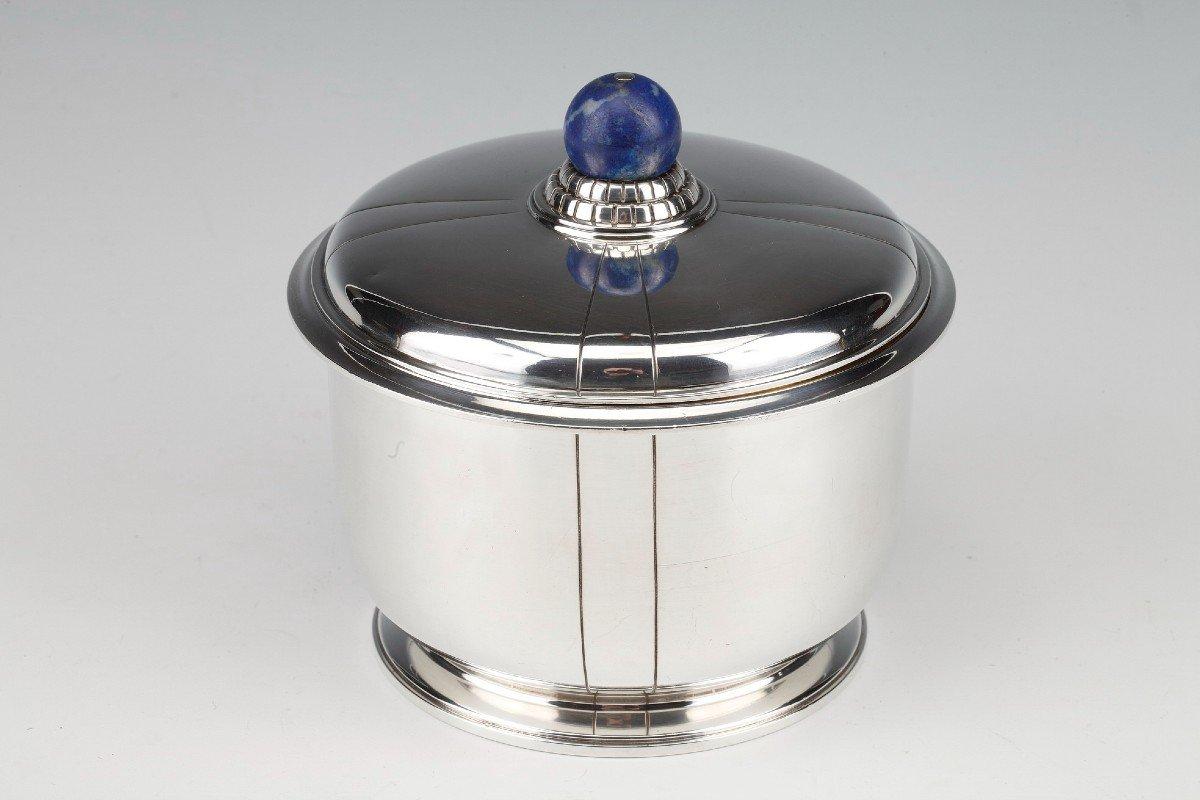 Cylindrical covered pot, forming a biscuit tin, resting on a circular base with a constriction. Silver proof.

The lid and the perimeter with fine engraved line patterns, the original lapis lazuli spherical socket is placed on a terrace in grooved