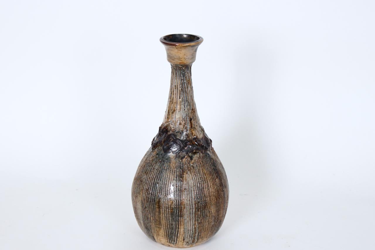 Slender Jean Hastings handcrafted glazed ceramic vase. Featuring a lipped bottle form, vertically heavily striated detail, Camel, Beige, Cocoa, Brown coloration and White translucent over glaze enhanced with hand applied glazed clay decoration. 3D