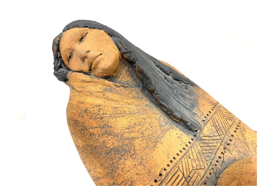 Scultura in gres che raffigura una figura femminile indiana sdraiata. La donna, a piedi nudi, indossa uno scialle con un intricato disegno traforato. La donna è in posizione reclinata con le braccia avvolte intorno al ginocchio. La scultura è
