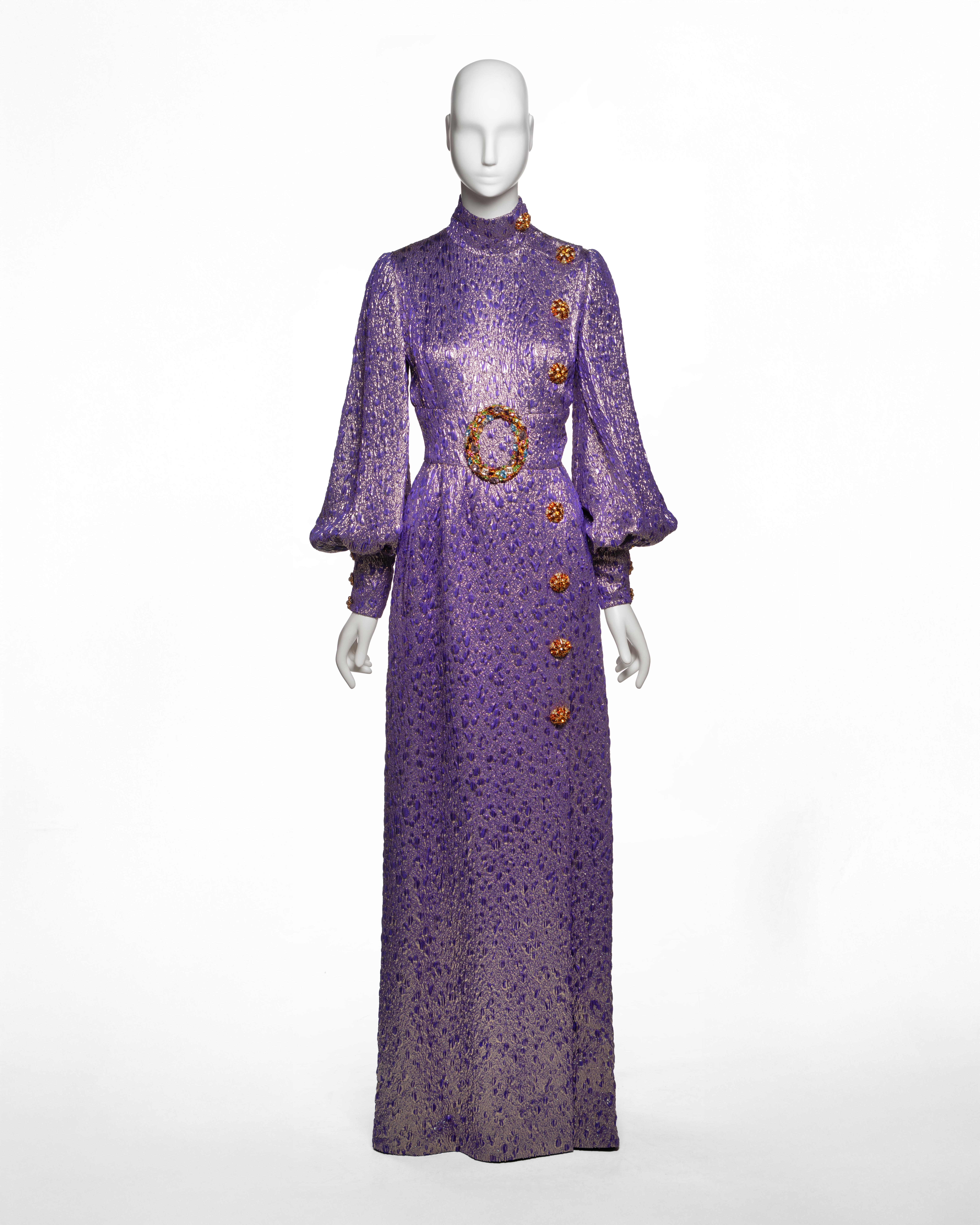 Este vestido de Alta Costura Jean-Louis Scherrer, diseñado por Stéphane Rolland y presentado como look nº 27 de la colección Otoño-Invierno 2005, está confeccionado en un brocado de lúrex púrpura y plateado con un tejido jacquard dimensional. La
