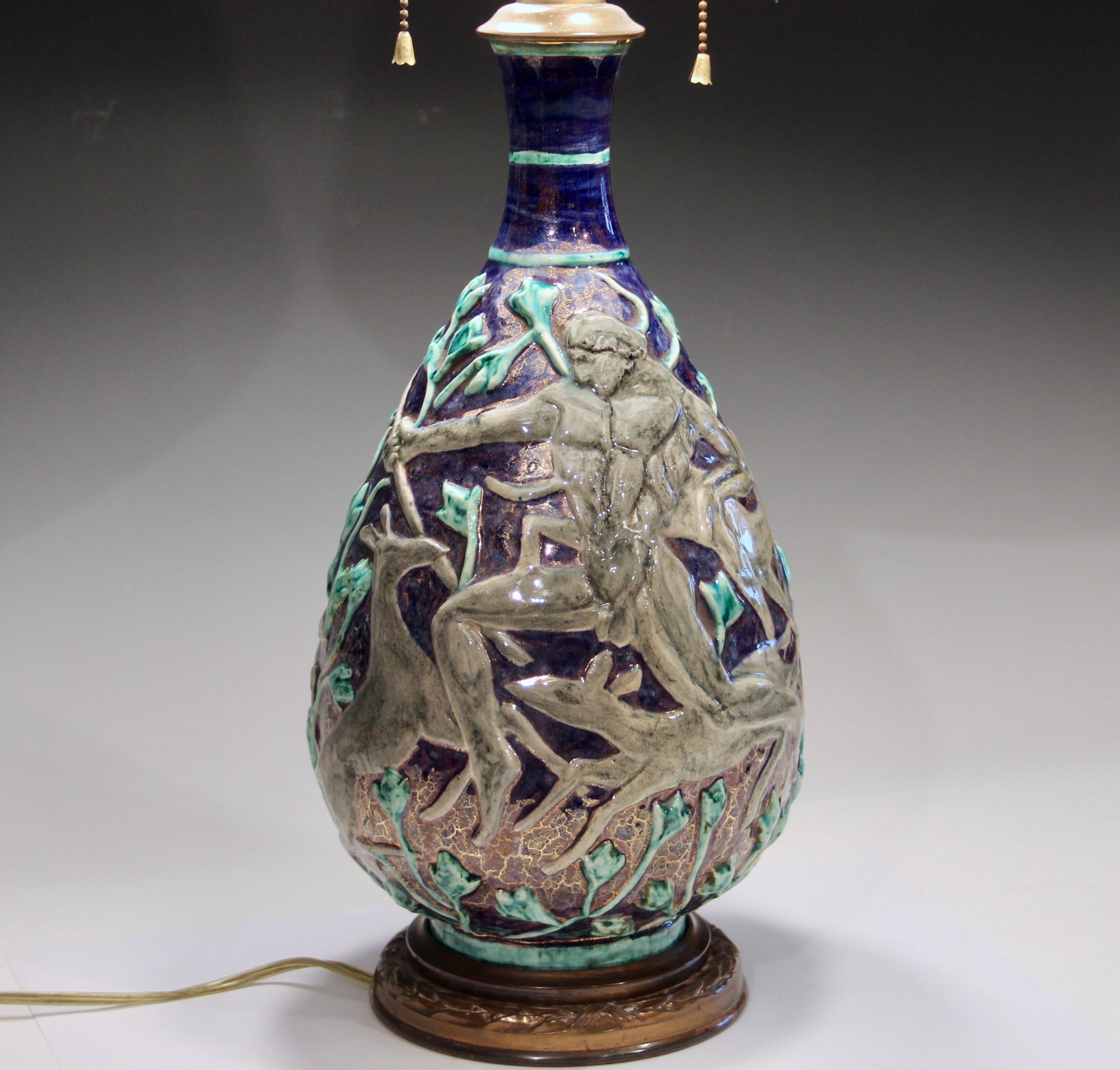Vintage Jean Mayodon Art Deco pottery lamp decorated with deer and hunters amidst a gilt blue and green forest. Circa 1920's. Nice quality brass hardware with mellowed patina. Double S socket cluster with smooth pulls and shade riser. Impressed