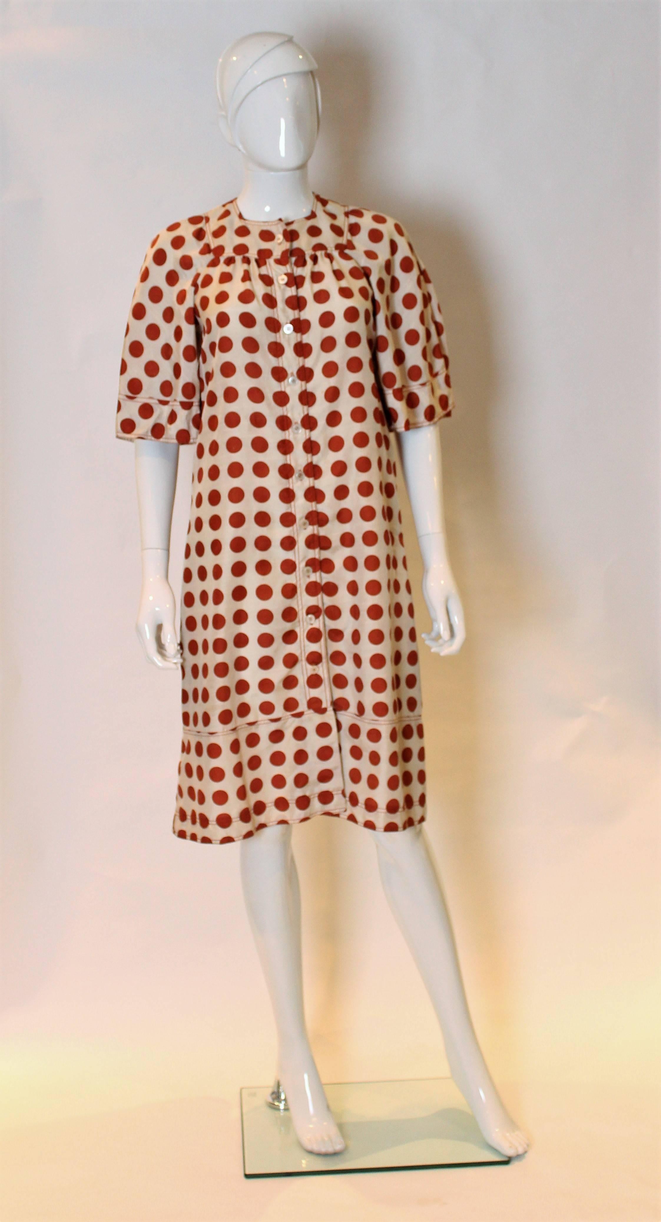A cute dress by Jean Muir, made for Harrods, London. The dress is made of sik and has an ivory background with brown polka dots. It has short angel like sleeves, a button through front, and is slightly flared at the hem. There are belt loops,but