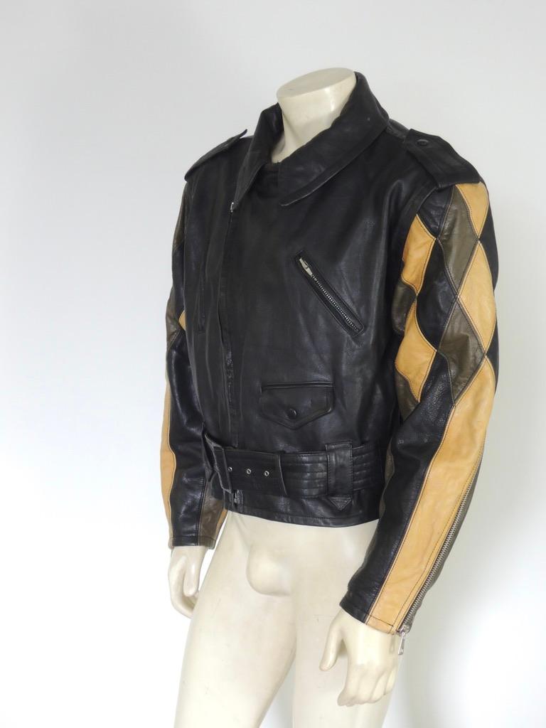 A vintage Jean Paul Gaultier black leather biker jacket with padded shoulders and a tapered cut. The shoulders have snap epaulettes. The sleeves of the jacket show a Harlequin style diamond patchwork design in black, brown, and tan leather. Starting