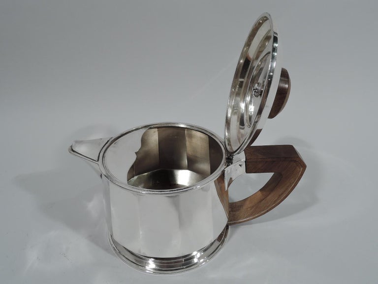 French Art Deco 950 silver teapot, circa 1930. Faceted drum on round and stepped foot. Covered “fish mouth” v-spout. Wood scroll-bracket handle. Cover hinged and raised with round and stepped finial; finial top has silver circle engraved with block