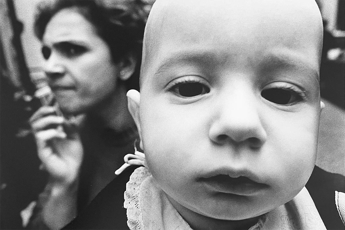 Naples #287 von Jed Fielding ist ein Silbergelatineabzug. Dieses Foto zeigt ein Babygesicht, das den Betrachter anschaut, während im Hintergrund eine Frau am Telefon vorbeigeht. Das Bildformat ist 12 11/16 x 18 15/16 Zoll, das Papierformat ist 16 x