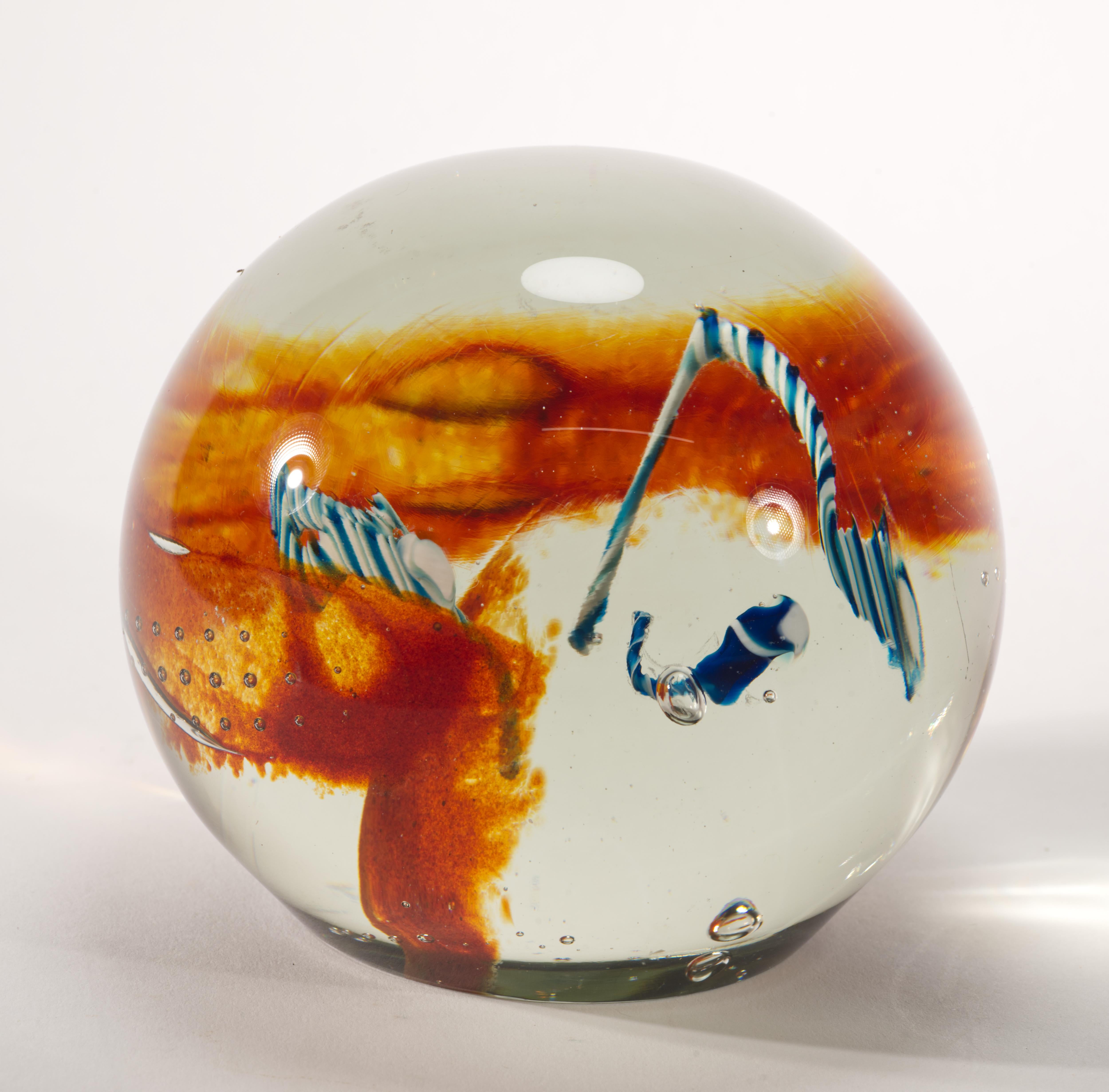 Massive, heavy paperweight features complex asymmetrical fused glass center, accentuated by groups of controlled bubbles. Three free-form string structures of blue and white glass are surrounded by veils of red pigments and controlled bubbles and