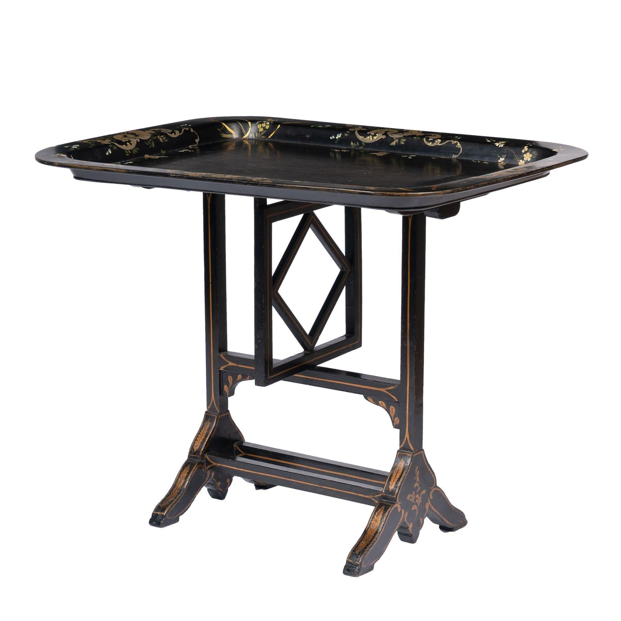 Rectangular paper mache tea tray table with a flat rim and rounded corners. The tray has a hand painted scroll work & floral border decoration and is mounted to a Jennings & Bettridge swivel brace ebonized wood stand with gilt stencil