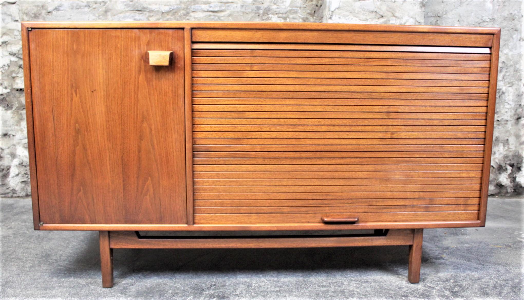 This small walnut credenza is done by the noted designer, Jens Risom and made in the United States in circa 1965 in the period Mid-Century Modern style. The credenza is done in dark walnut with both a swing door and tambour door on the front with