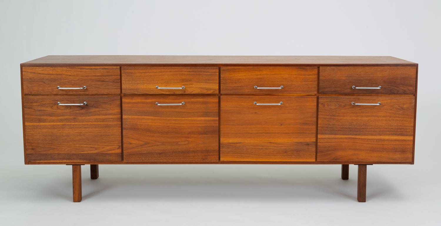 A walnut case piece from Jens Risom design has a low profile and sits atop four squared wooden legs. The credenza accommodates eight storage drawers, in two rows of four. The upper drawers are shallow; the far left drawer bears the Risom Design
