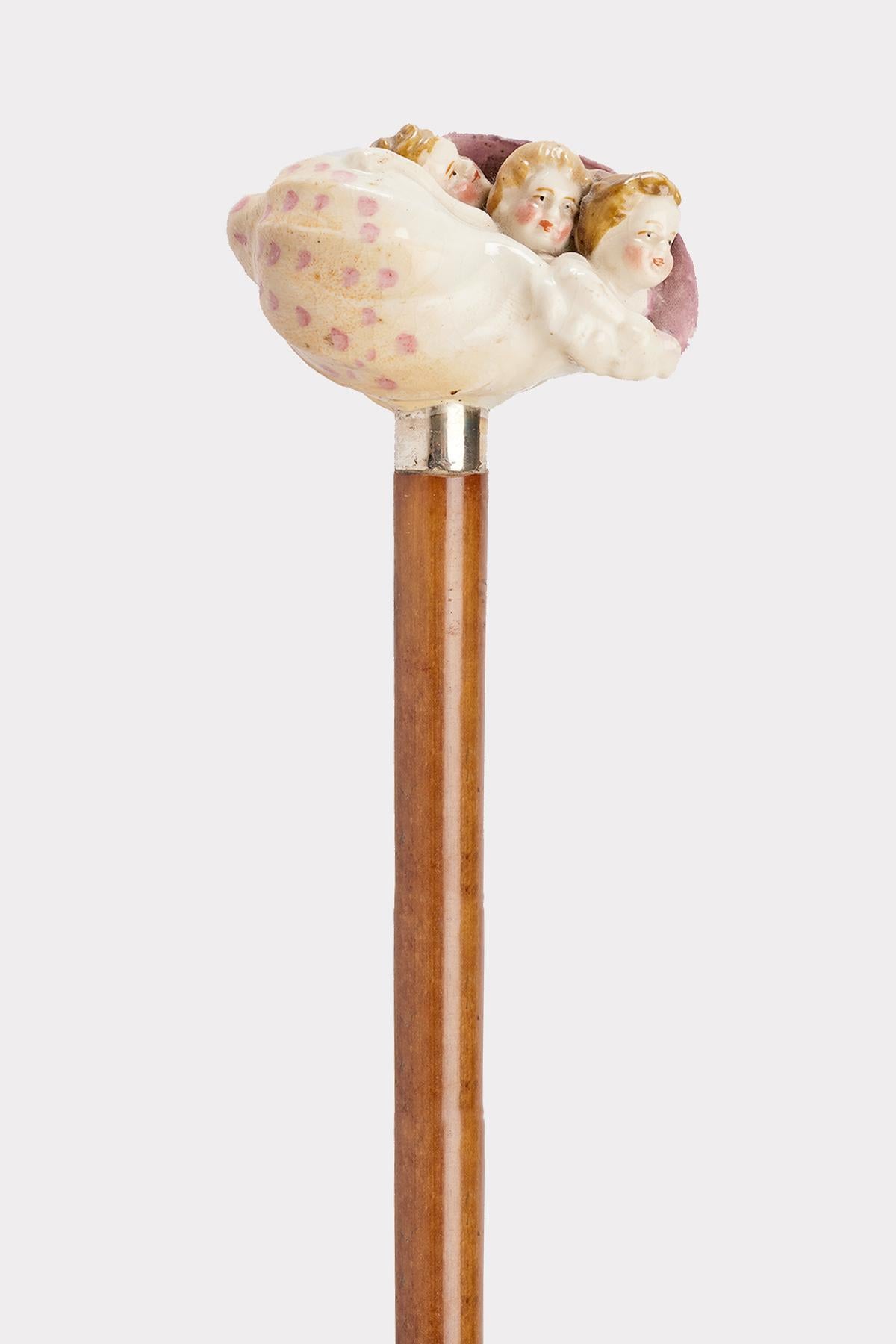 Canne : poignée en porcelaine peinte, manufacture de Capodimonte, représentant un coquillage Cyprea, avec trois têtes d'enfants émergeant de la cochlée du coquillage. Bande argentée. Manche en bois de rotin. Virole en métal. Naples, Italie vers 1880.