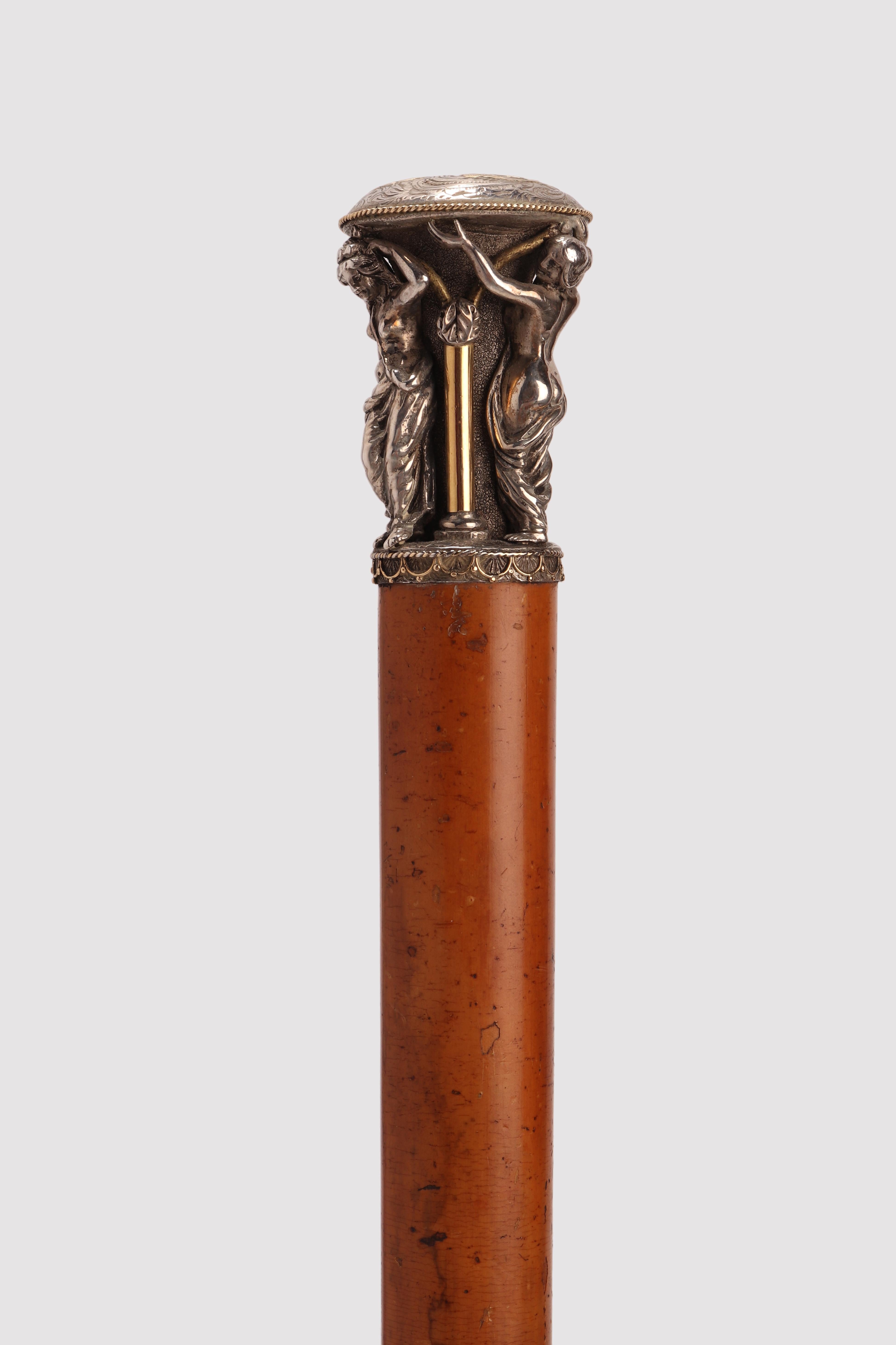 Walking stick. The handle is craftsmanship, made out of silver and vermeil. The ring is silver, chiseled, with golden arches and small spheres. A golden filigree cord underlines the beginning of the handle itself. It shows the three Graces,