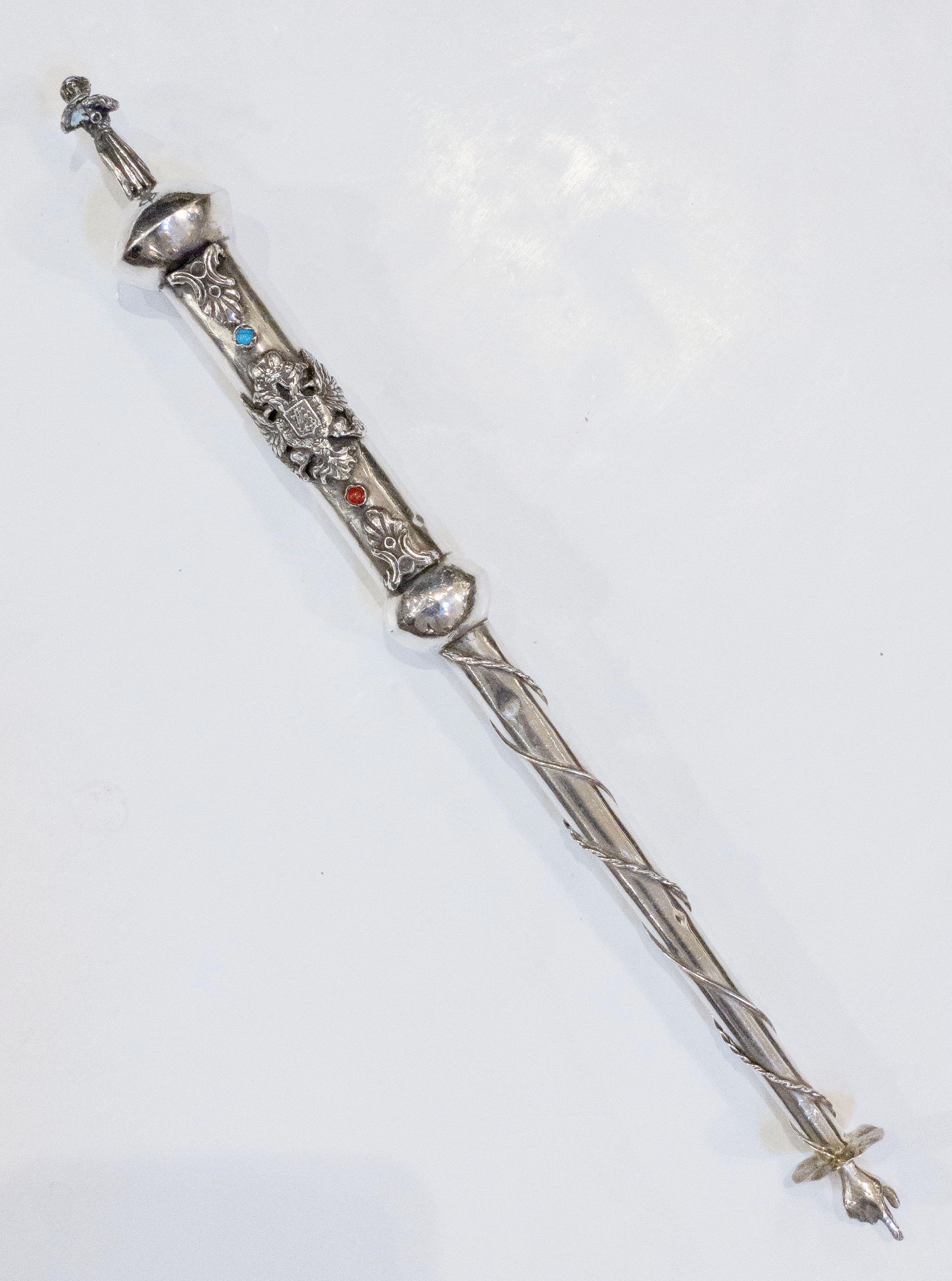 A fine Jewish yad or torah pointer of silver featuring a robed man playing a wind instrument on the handle end, with a traditional pointed finger hand on the other end. With decorative blue and red stones. A handsome find for the Judaica