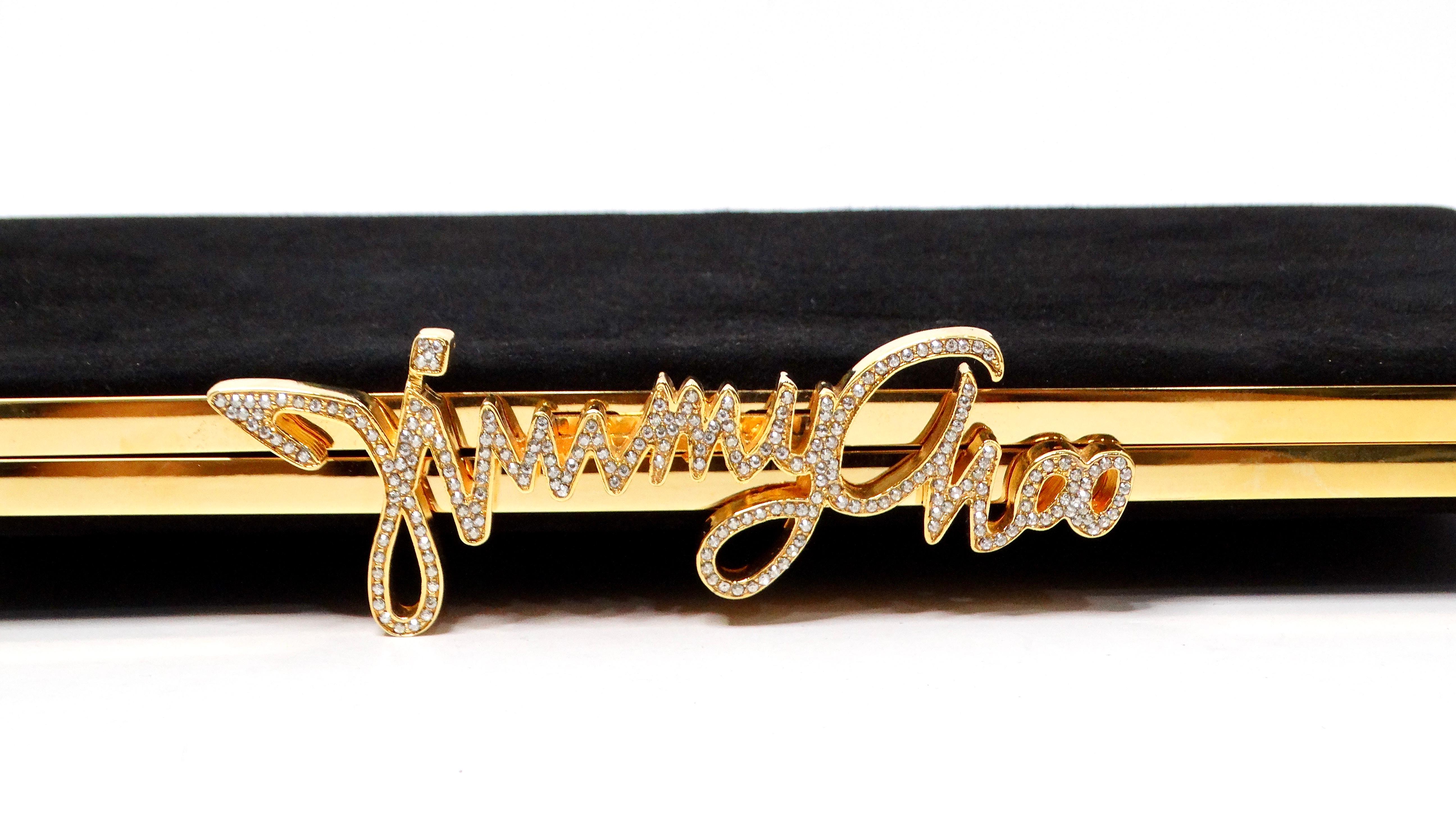 Te presentamos el Bolso con Logotipo de Cristal Celeste de Ante Negro de Jimmy Choo, epítome del lujo y la elegancia. Este bolso de mano rectangular, confeccionado en suntuoso ante negro, está adornado con llamativos herrajes dorados que realzan a