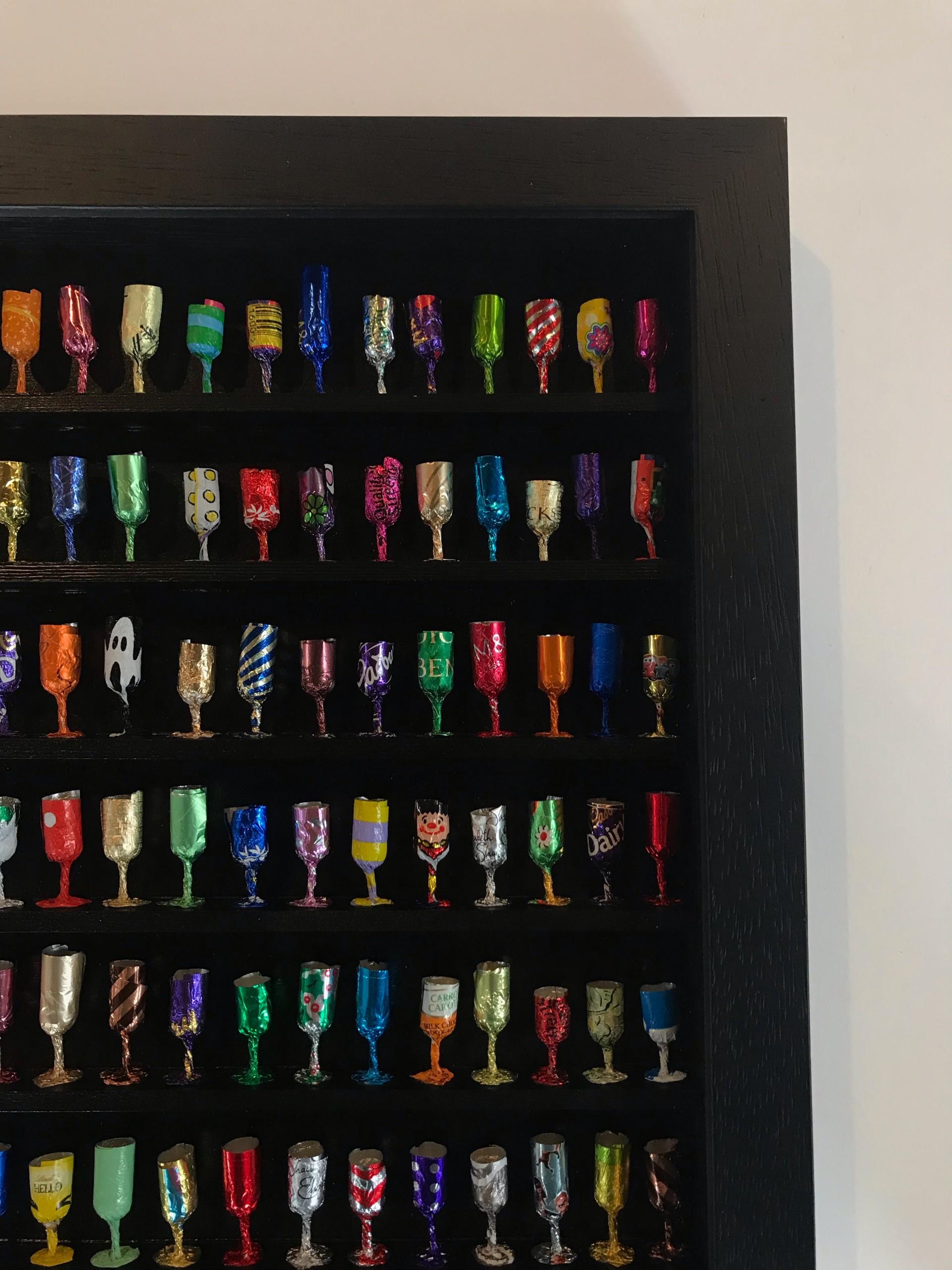 Goblets XXV is a signed original wall sculpture made from sweet wrappers by Joanne Tinker of a selection of colourful wine goblets on a black shelved frame.
Joanne Tinker wall sculptures available at Wychwood Art. Joanne Tinker seeks to transform