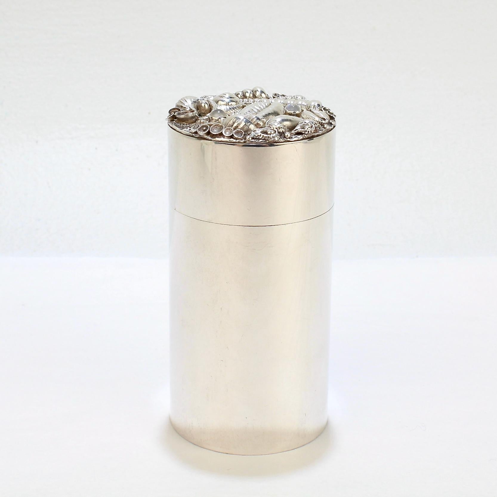 A very fine cylindrical sterling silver box by Jocelyn Burton.

The box's lid is richly encrusted with miniature silver sea shells and bezel set with 2 moonstone cabochon gemstones. 

The sides of the box are thick-gauged and the lid slides on