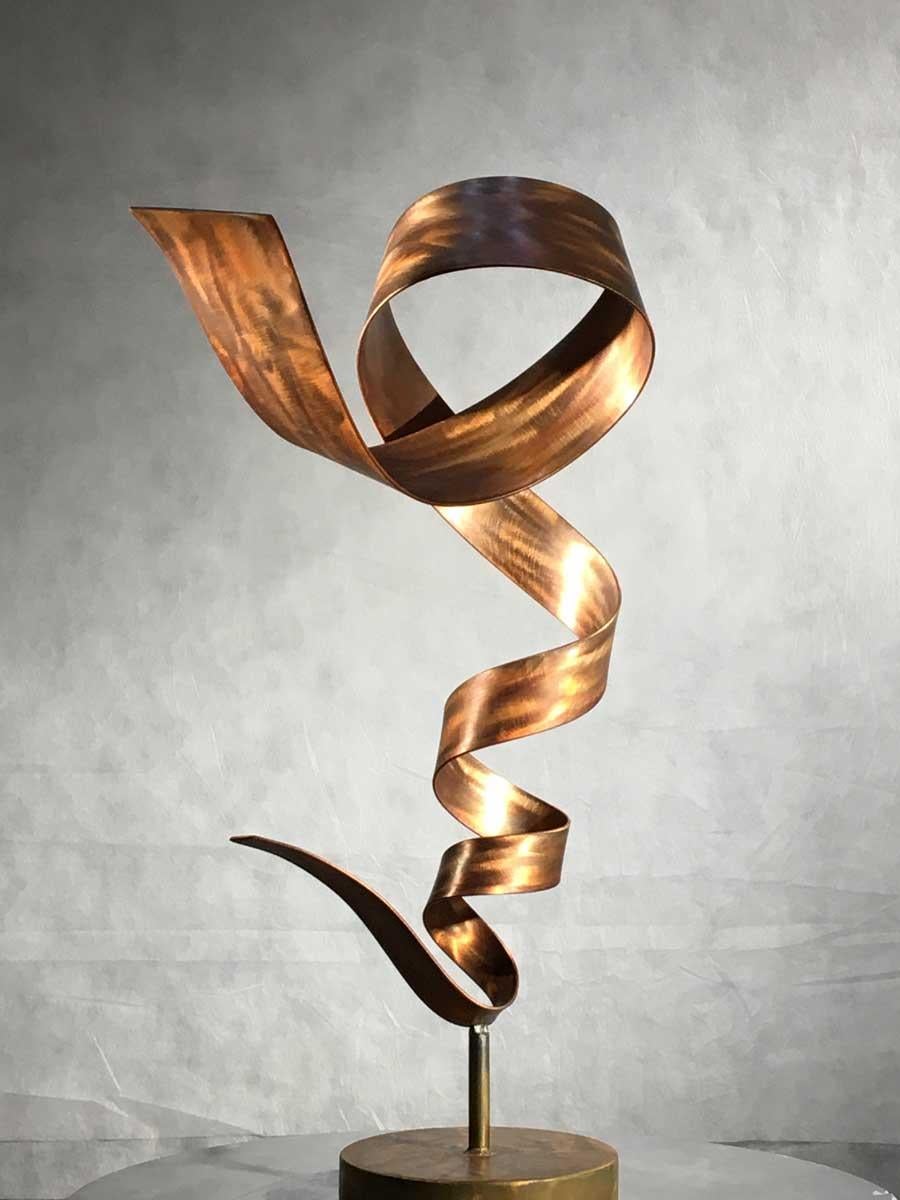 This mid-sized abstract contemporary sculpture by Joe Sorge is made with steel, orange dye, and wax. The warm-toned piece features a strip of steel metal that twists and turns in a flowing, spiraling movement. It casts unique shadows on its