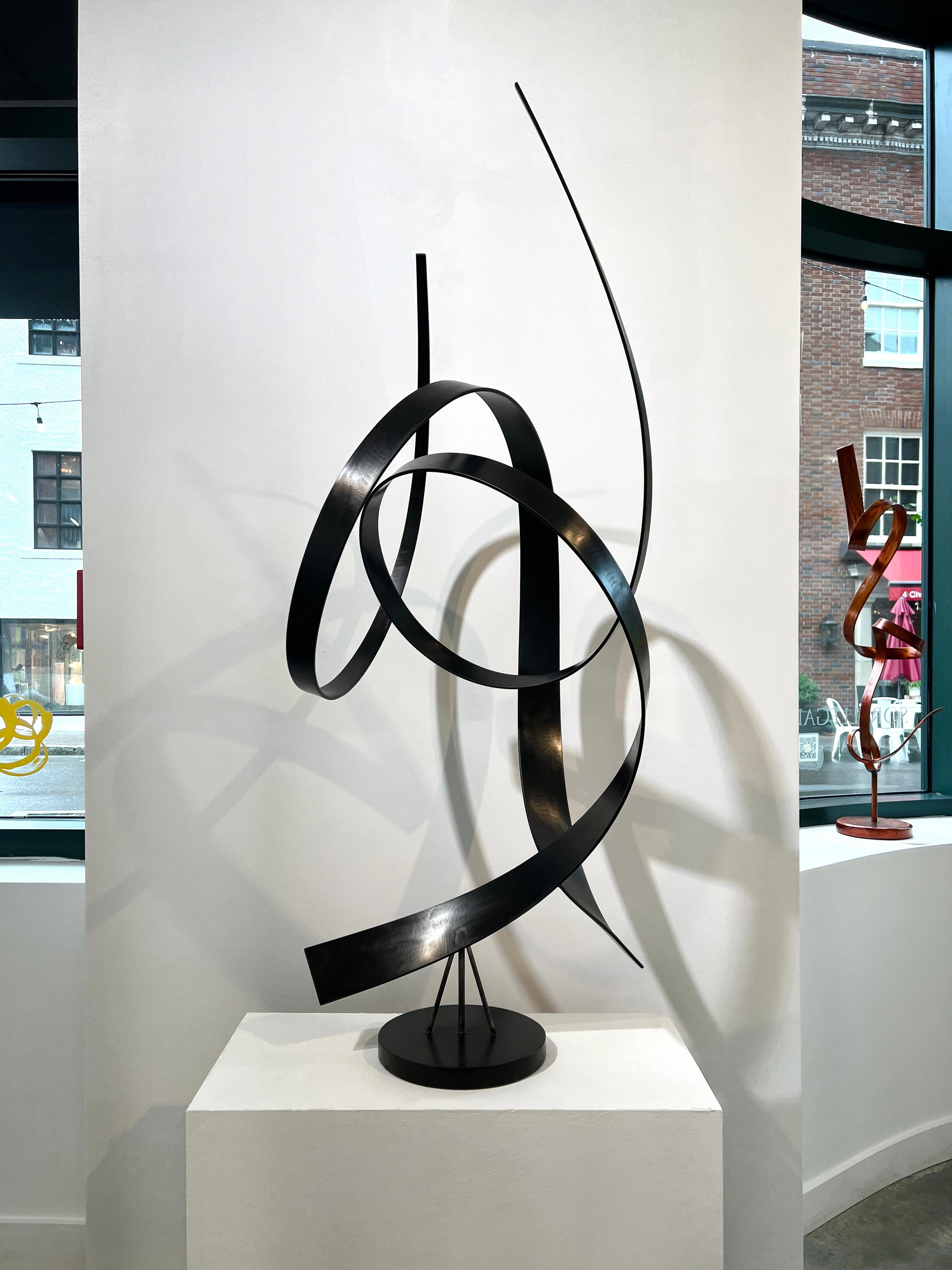 This large, abstract sculpture by Joe Sorge is made with steel and black dye. Two strips of steel curl upward from a round, circular base, twisting through and around one another. The sculpture casts unique and intricate shadows on its surrounding