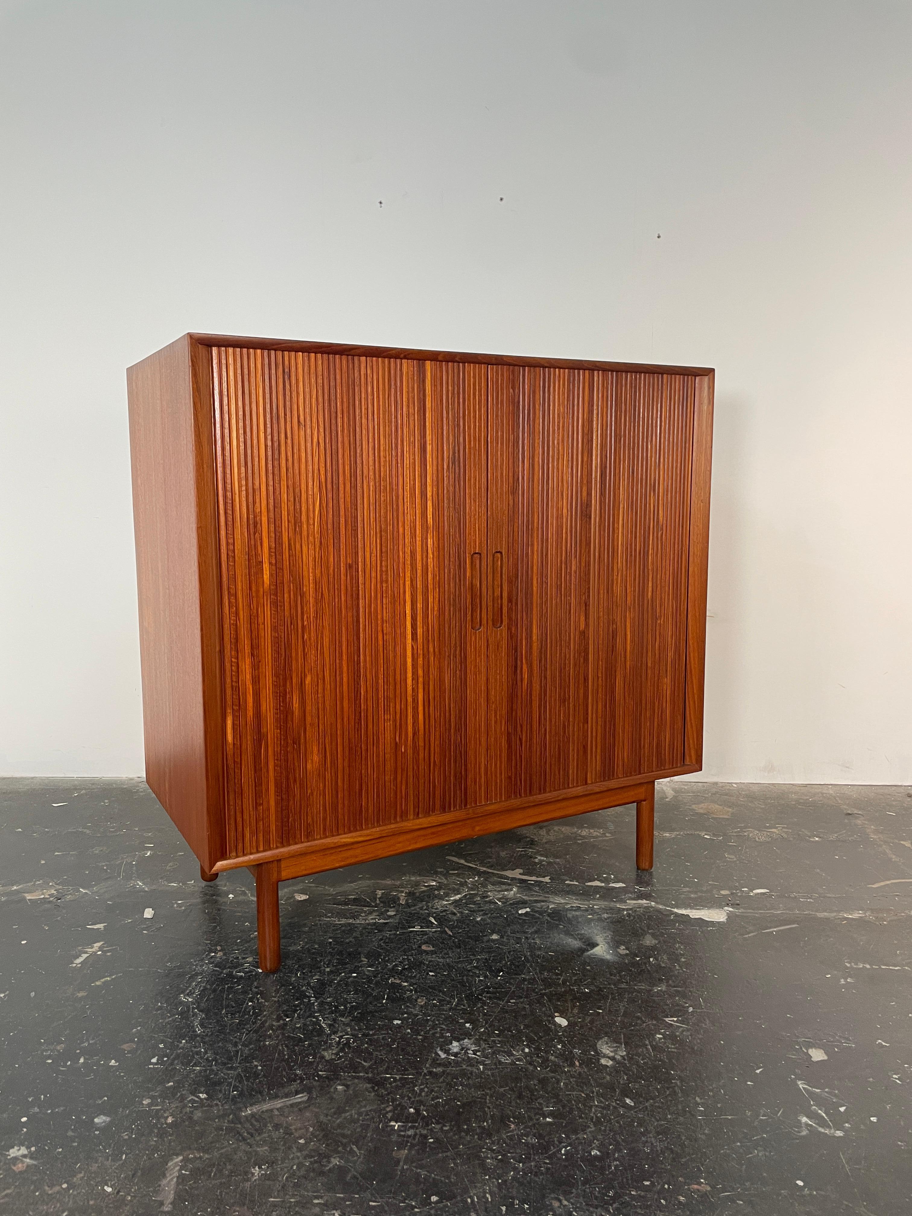 Raro armario alto con puerta de tambor Johannes Aasbjerg de teca maciza. Este armario con puerta de persiana brilla en persona, la madera de teca es preciosa y de un color rịch. Esta pieza también presenta uniones dentadas y un borde biselado a