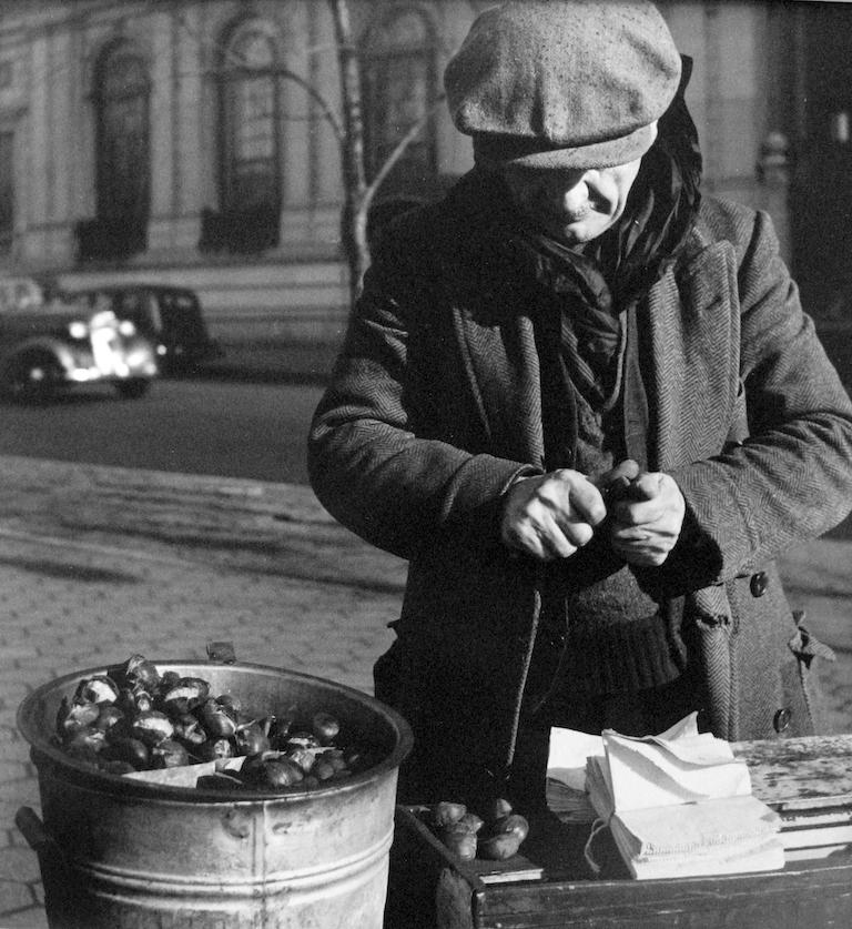 Chestnut Vendor, Depression von John Albok zeigt einen Mann, der auf der Straße Kastanien verkauft. Der Mann schaut auf seine Hände hinunter, während er die Kastanie aufbricht. Zu seiner Linken steht eine Schale mit Kastanien, und unter ihm ein