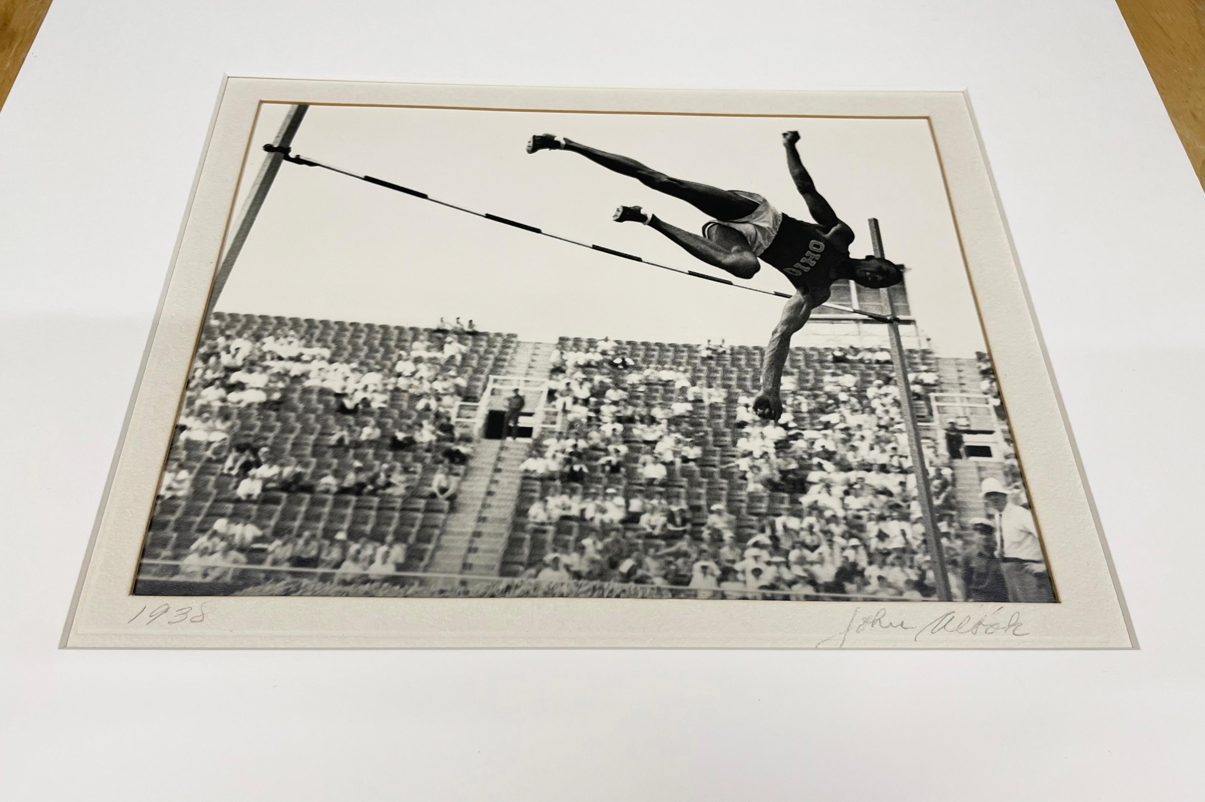 Randall's Island Worker's Sports Day (Track and Field) by John Albok features an athletic man performing a pole vault, frozen in space as he begins the descent down towards the mat. Stadium seats are visible behind the athlete, with the crowd seated