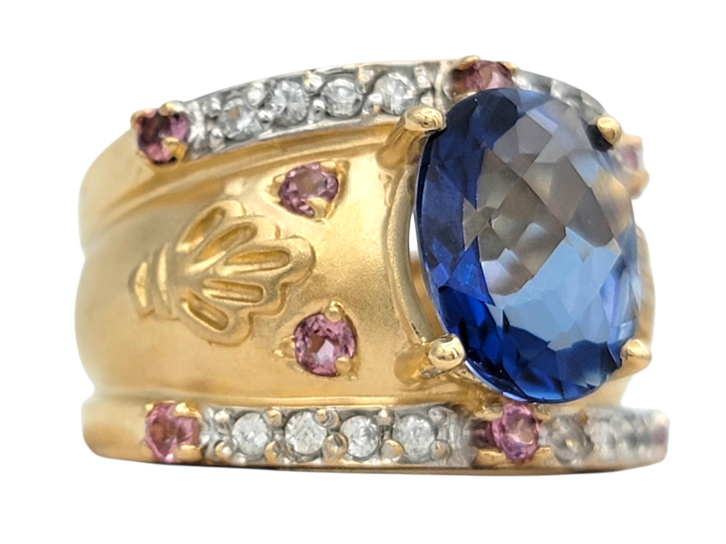 This striking John C. Rinker ring features a bold blue topaz center stone that immediately draws the eye with its vivid color and dimensional checkerboard reflections. Surrounding the center, a row of soft pink accents adds a delicate contrast that