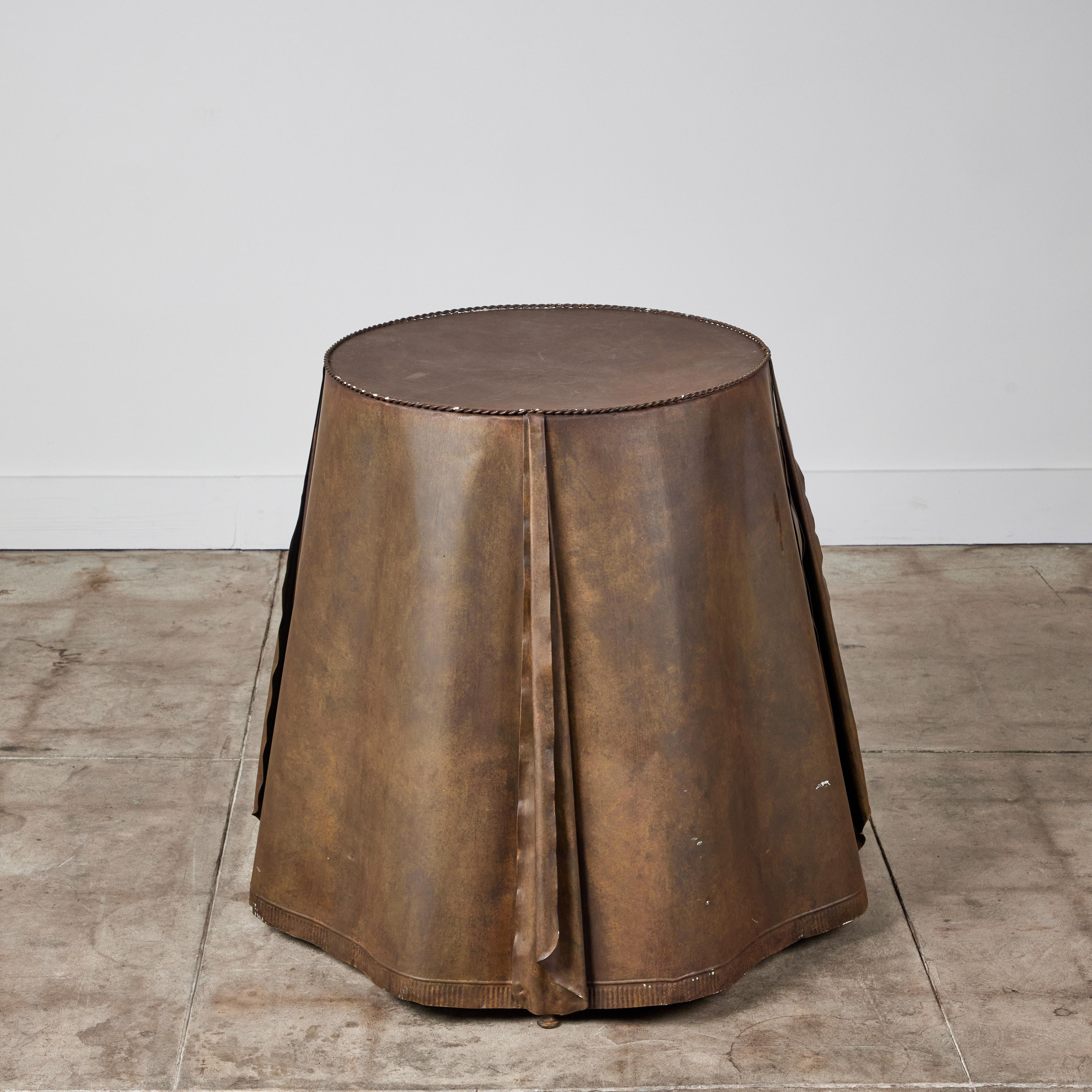Steel side table, created in the style of American designer John Dickinson. The round table imitates draped table cloth, with sculptured pleats and folds, the surface is finished with a painted faux brass patina.

Stamped - Made in