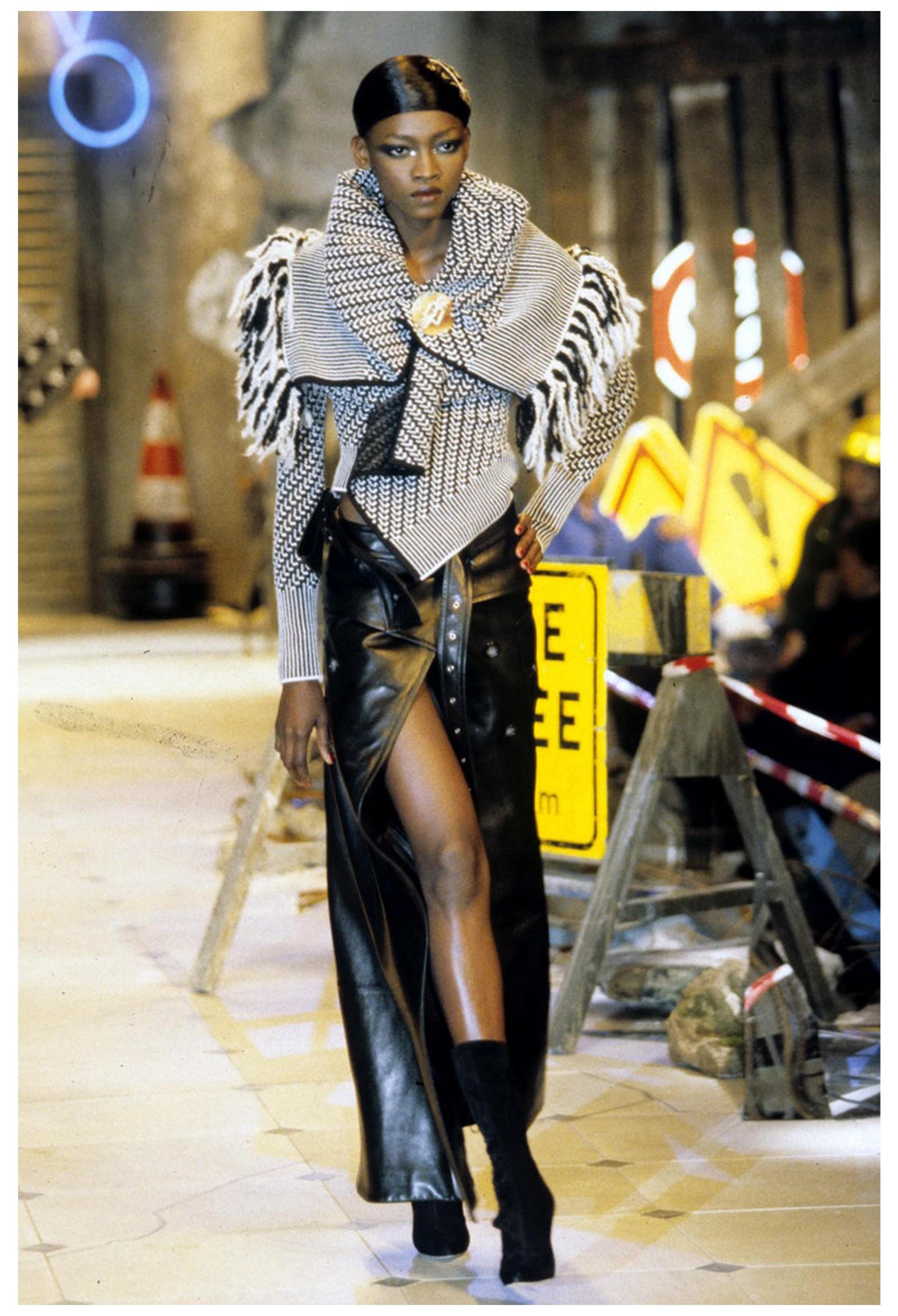 Resurrection Vintage se complace en ofrecer una falda trench vintage de lana negra de John Galliano de la colección otoño-invierno 1999. La falda lleva botones asimétricos de doble botonadura en la parte delantera, una abertura alta en la pierna, un