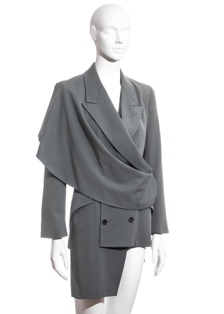 ▪ John Galliano dusty teal double breasted blazer jacket
▪ 100% Wool
▪ 100% Silk lining 
▪ Capelet panel to one shoulder fastening through a belt loop on opposite hip
▪ Asymmetric hemline 
▪ UK 8 
▪ Fall-Winter 1988

* Please note that this piece is