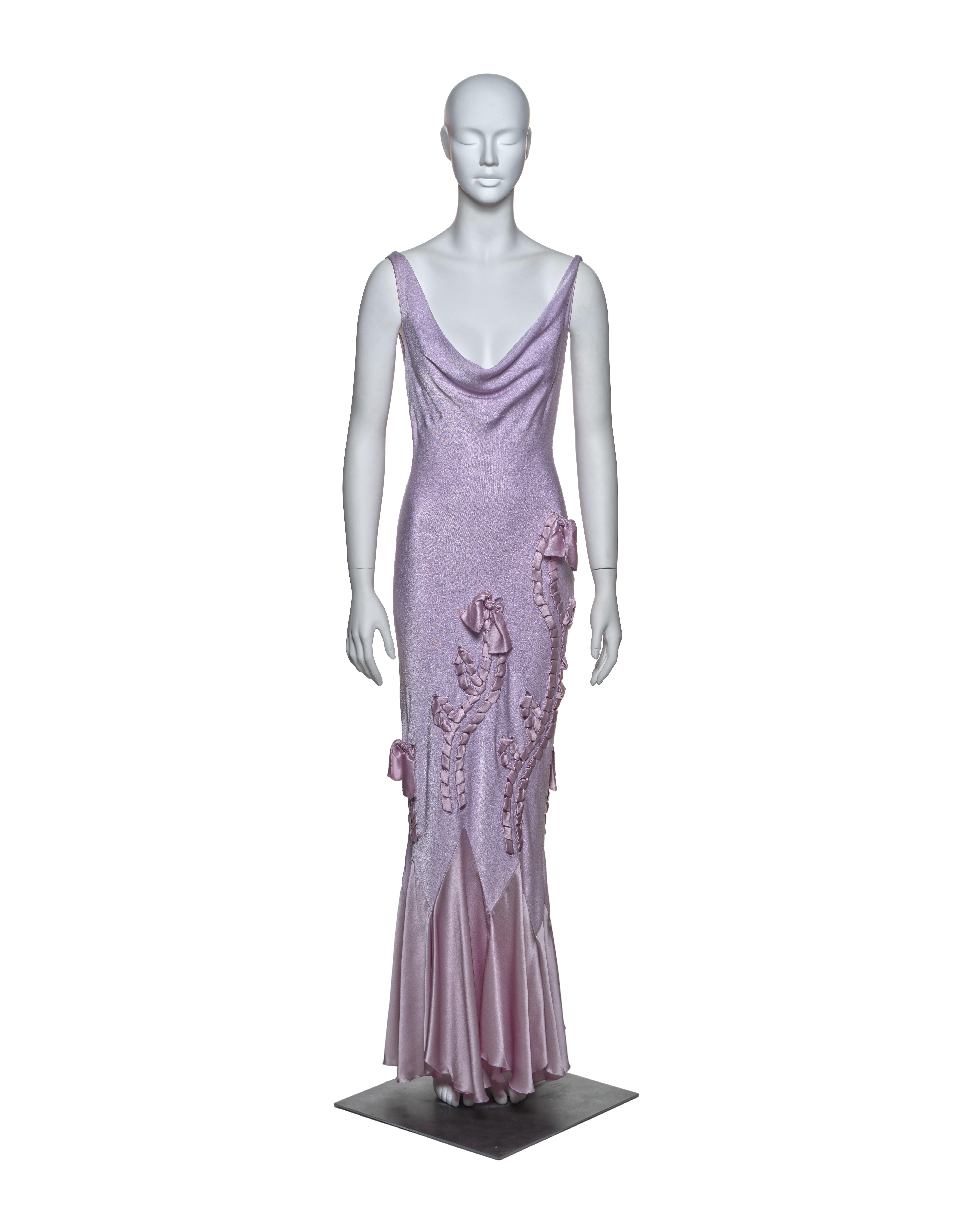 This John Galliano bias-cut satin gown in soft lavender features a draped cowl neckline and an open back. Satin ribbon appliqués run along the skirt, adorned with bows for added detail. The bias cut allows the fabric to fall fluidly, accentuating