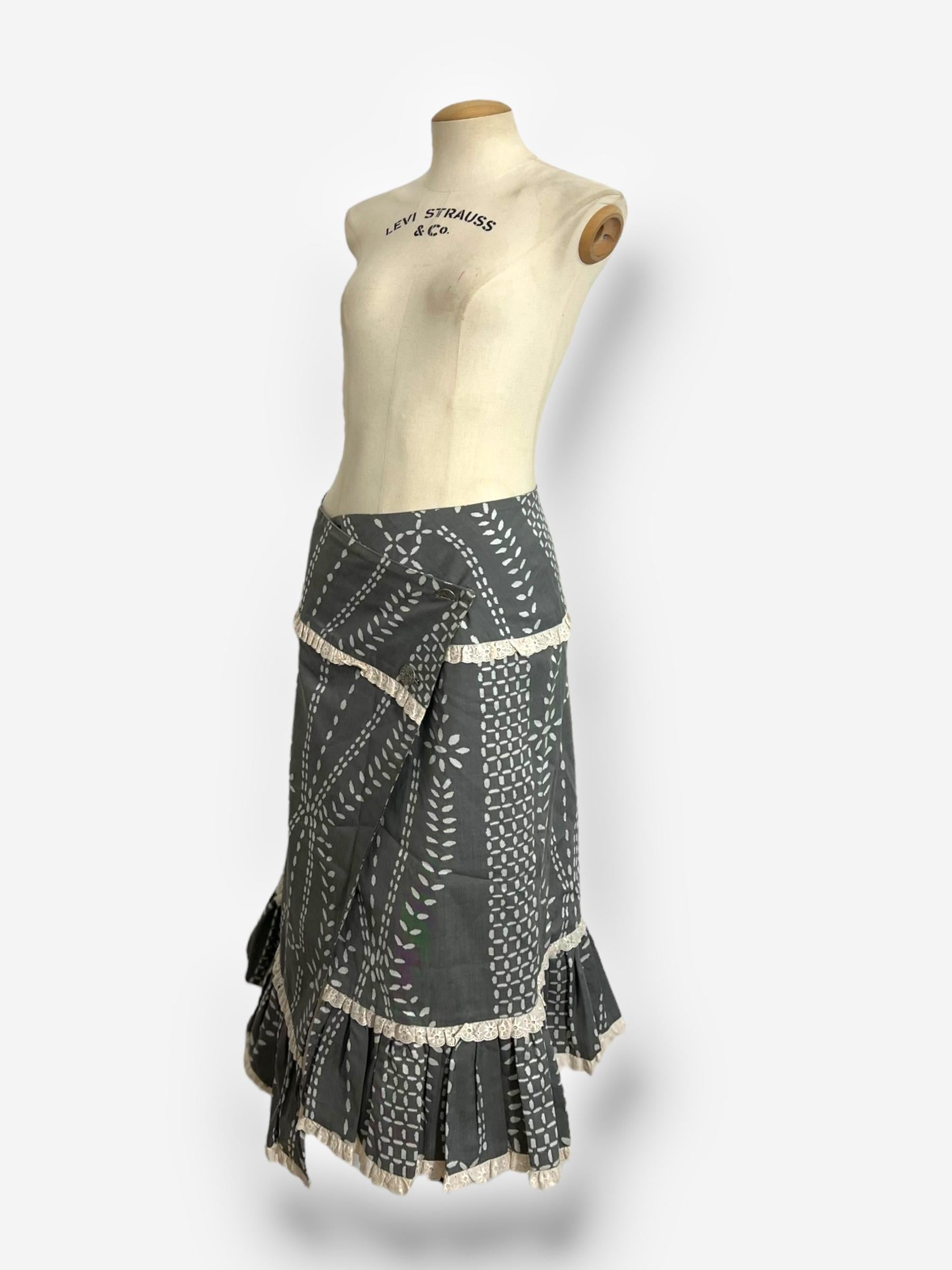 A romantic and deconstructed wrap-style cotton skirt from John Galliano's Spring/Summer 2002 collection. This piece features:
* Delicate white folk-inspired print on a muted grey cotton base, creating a vintage, bohemian aesthetic.
* Lace trim