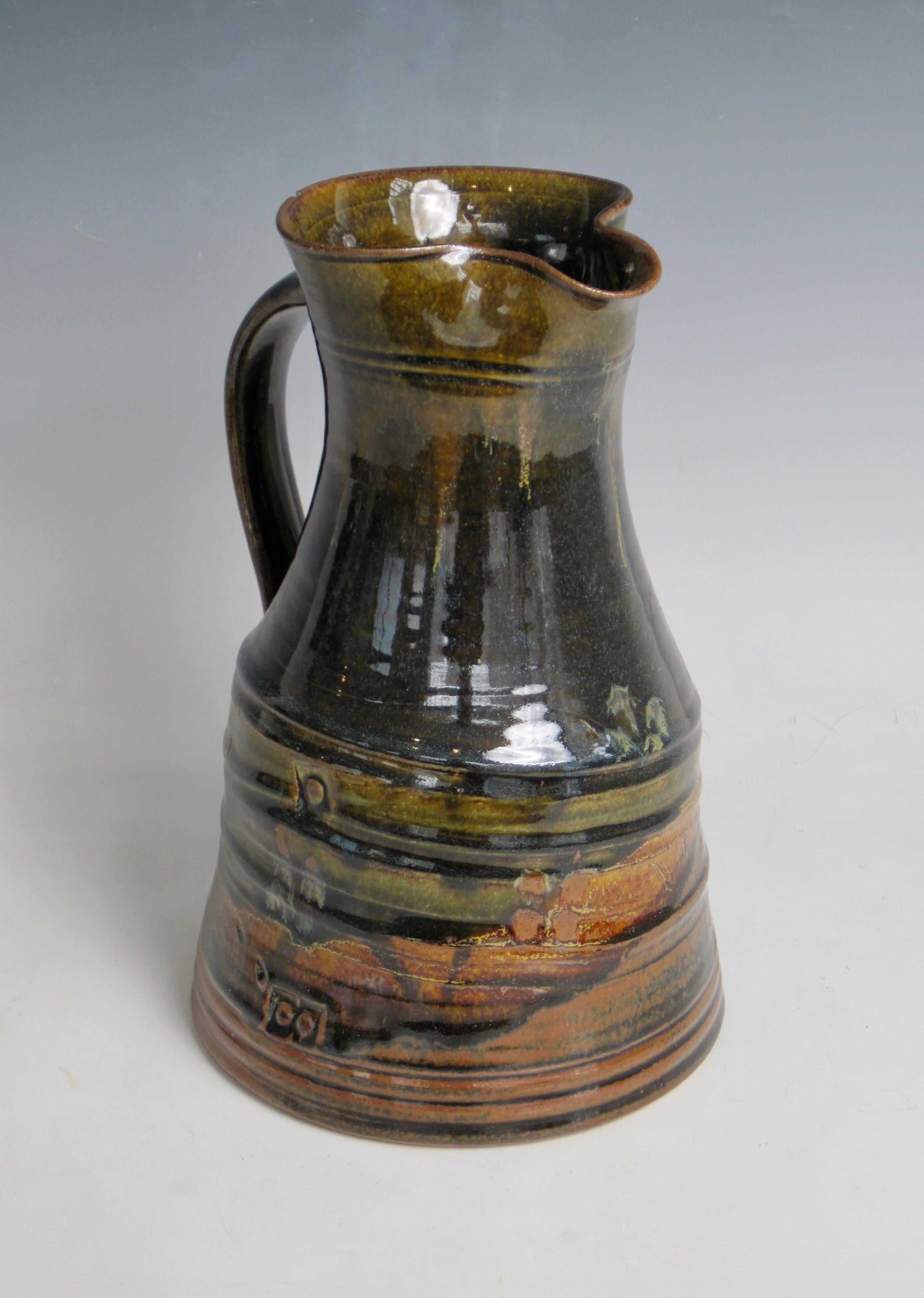 A traditional form pottery pitcher by John Glick in shades of brown, green and orange. Markings on sides and signed on underside.
John Glick is a people’s potter. In a career spanning over five decades, the ceramist has remained committed to the