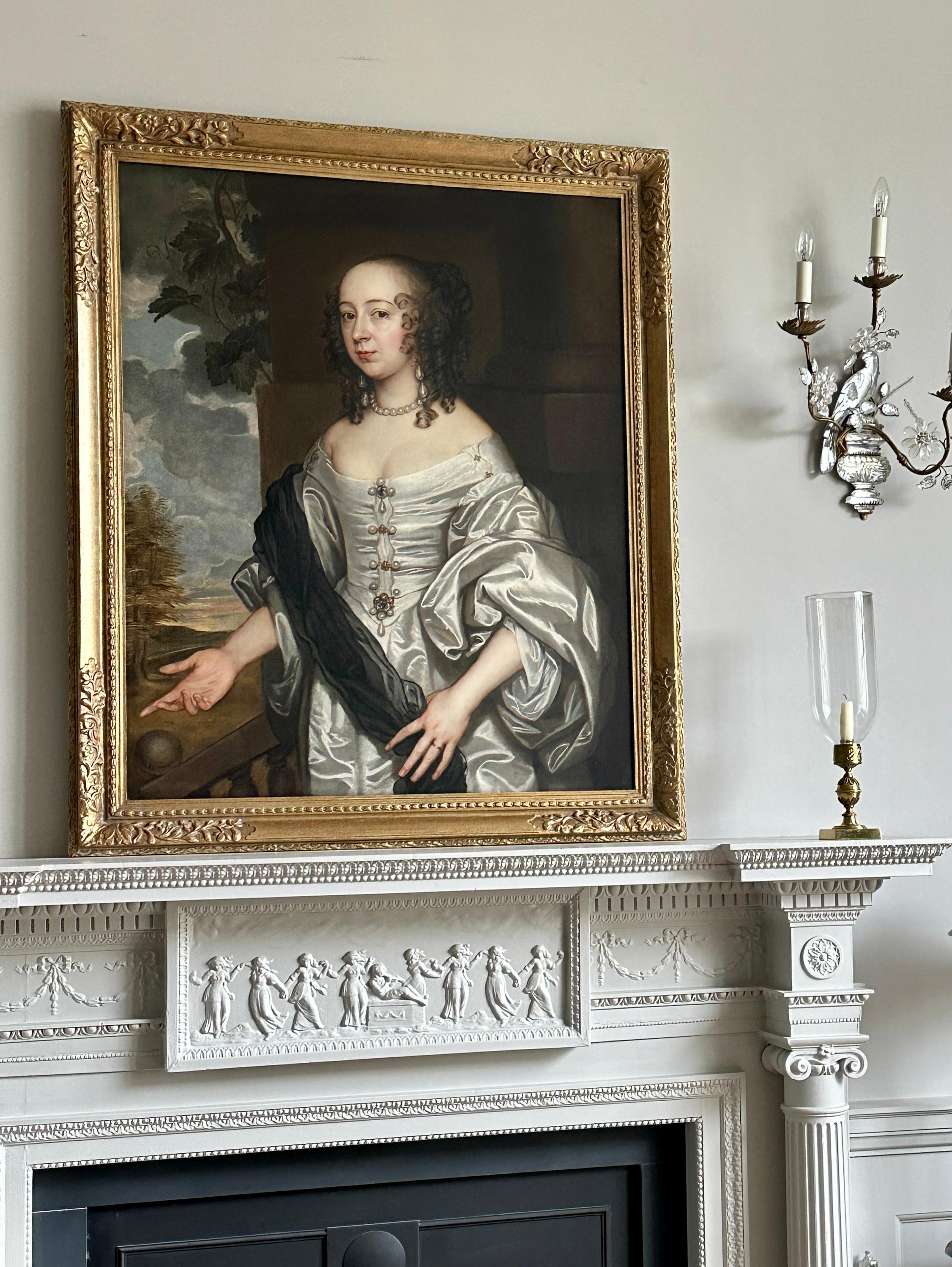 Portrait of a 17th century lady, three-quarter length, in an ivory silk gown standing on a garden terrace.
The lady, painted circa 1670, gestures with her hand towards the gardens and land beyond off her country estate, wearing a sumptuous ivory