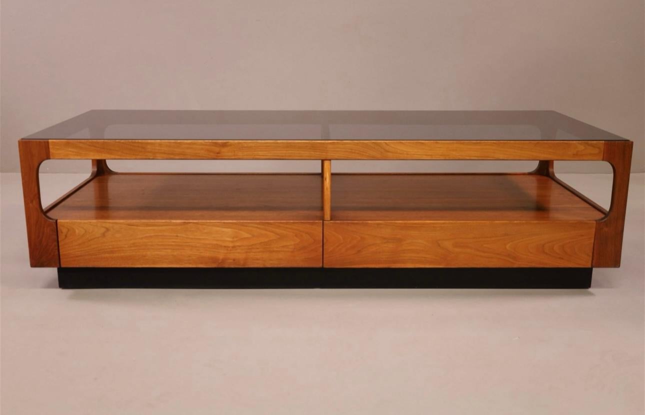 Une belle et élégante table basse à un tiroir conçue par John Keal pour Brown Saltman. Fabriqué en noyer, le meuble est doté d'un plateau en verre fumé.  Les dimensions sont de 60x24 pouces et la hauteur de 16,5 pouces.  Le veinage du bois est