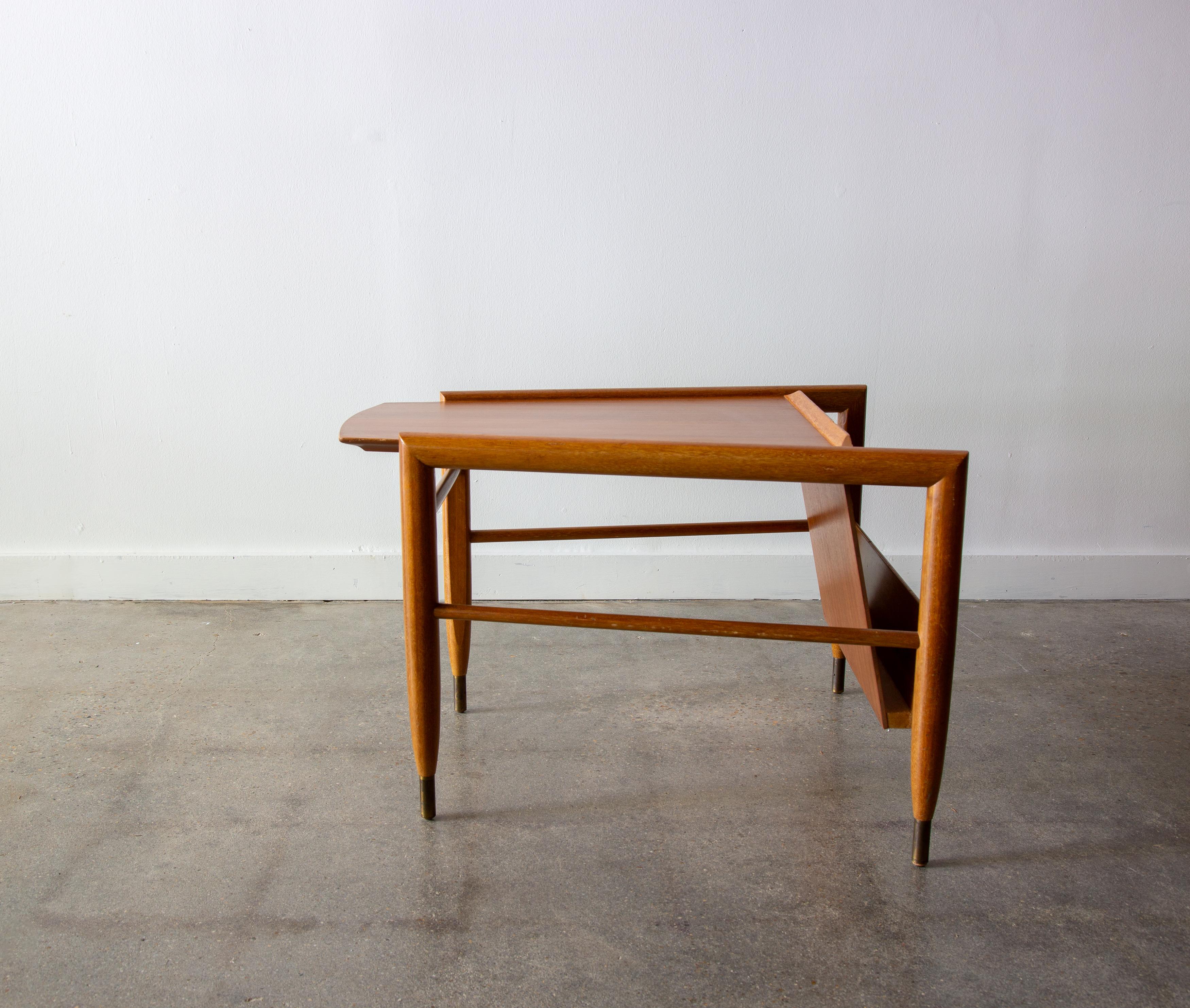 A mahogany wedge shaped table designed by John Keal for Brown Saltman.  The wedge shape allows it to nest between a pair of chairs. Tapered stiletto brass capped legs  A sleek floating magazine rack for ample storage and displaying your favorite