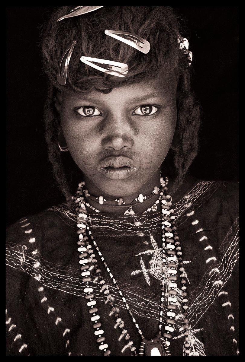 A young Wodaabe girl on route to local village market in Niger. She is dressed in the traditional dark cloth of the nomadic Wodaabe, which beautifully compliments the colourful beads and bright eyes of Wodaabe women. The hair clips are a modern