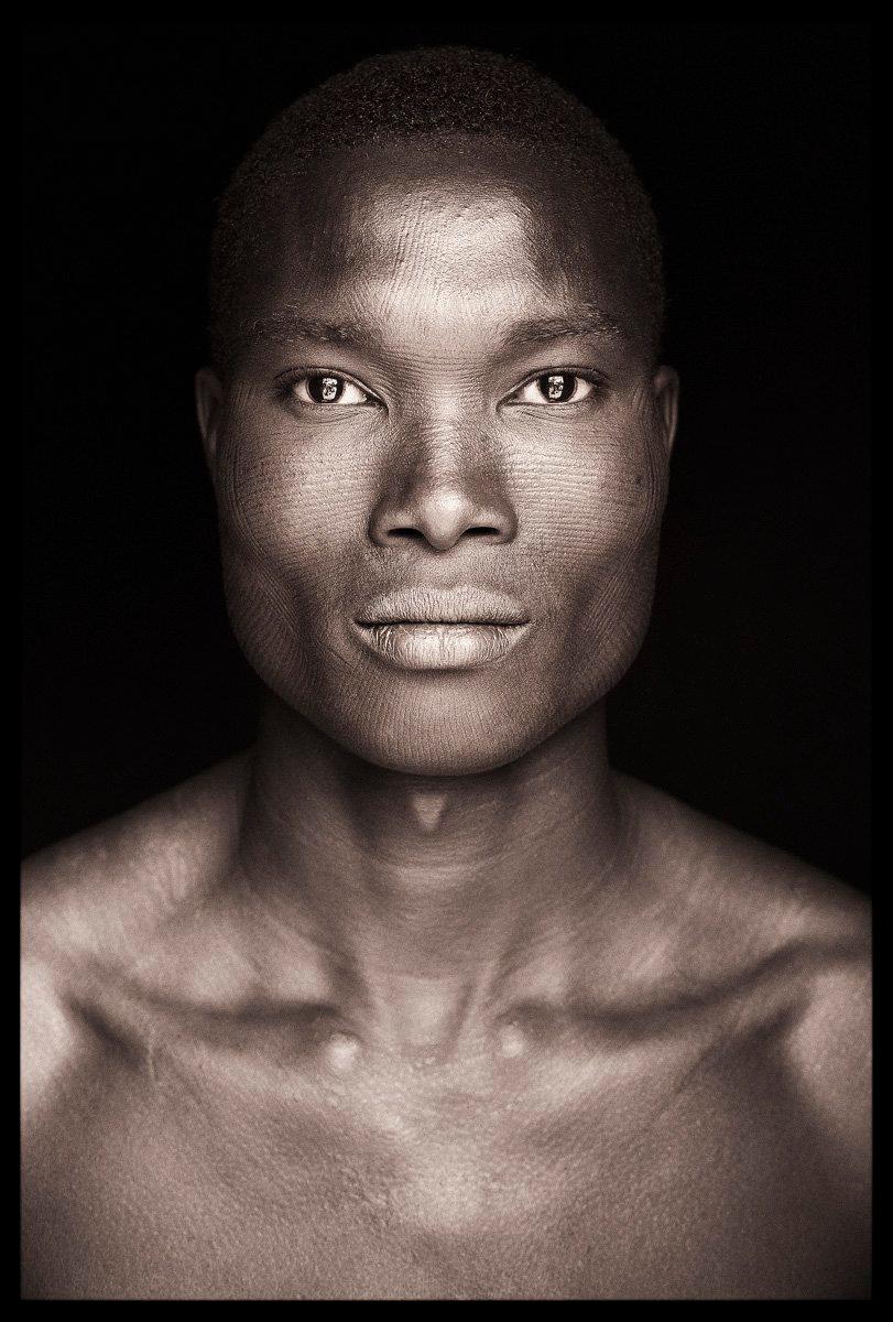 Ydaco stammt aus der Betammaribe-Kultur in Benin. Er lebt in einem kleinen Dorf in den Atakora-Bergen. Seine Wangenknochen bieten die perfekte Oberfläche für die wunderschön verschlungenen Skarifikationen seines Stammes und zeigen das Handwerk eines