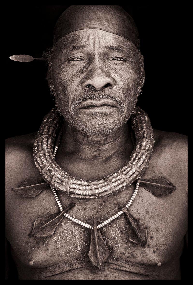 Dieser Himba-Älteste nahm an einer großen Begräbnisveranstaltung im westlichen Kaokoland zwischen der größten Stadt des Gebiets, Opuwo, und Otjitando teil. Die Gegend war flach und sehr trocken, und ein starker Wind peitschte ständig feinen,