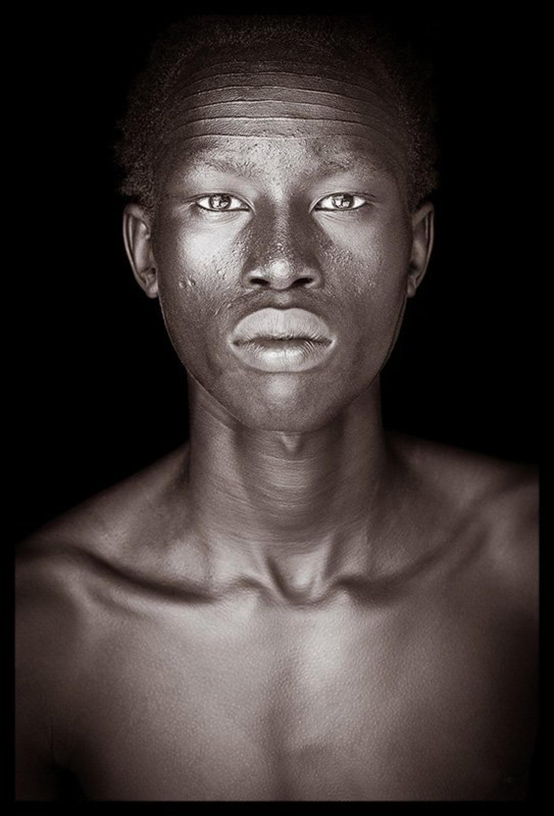 Ein junger Mann der Nuer-Kultur mit einer auffälligen Narbe auf der Stirn.  Das Foto wurde in Äthiopien aufgenommen, aber wie viele der Nuer wurde er durch den Konflikt aus seiner Heimat im Südsudan vertrieben.
John Kennys Arbeiten werden alle vor