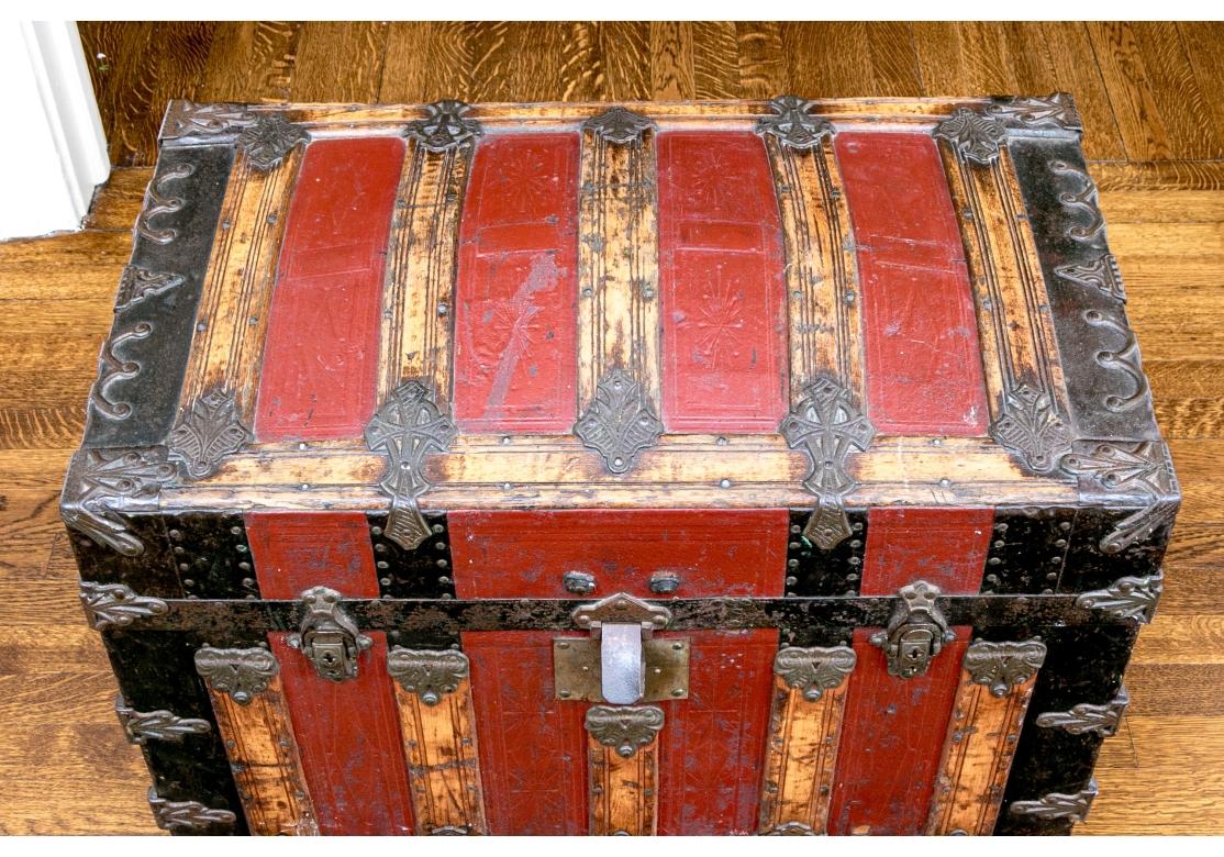 Une malle à vapeur colorée de John P. Shriner & Co., de forme bombée classique, avec des panneaux vibrants en cuir noir et rouge orangé, alternant avec des bandes de métal, des supports décoratifs et des garnitures en tête de clou autour des coins.