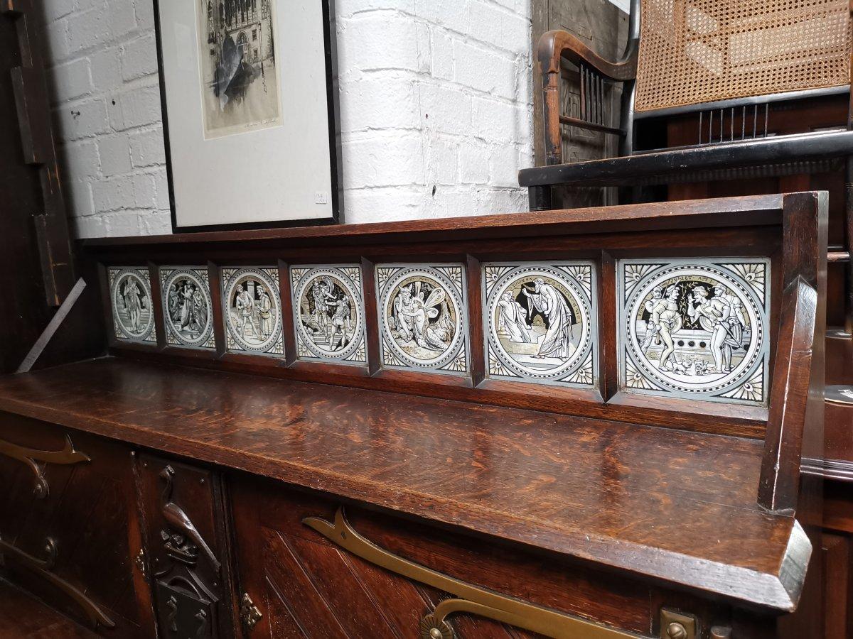 John Pollard Seddon, style of.
A Gothic Revival oak sideboard/buffet, with seven Minton Shakespeare series tiles, designed by John Moyr Smith to the top shelf and a carved armorial below flanked by a pair of upper cupboards and a pair of large