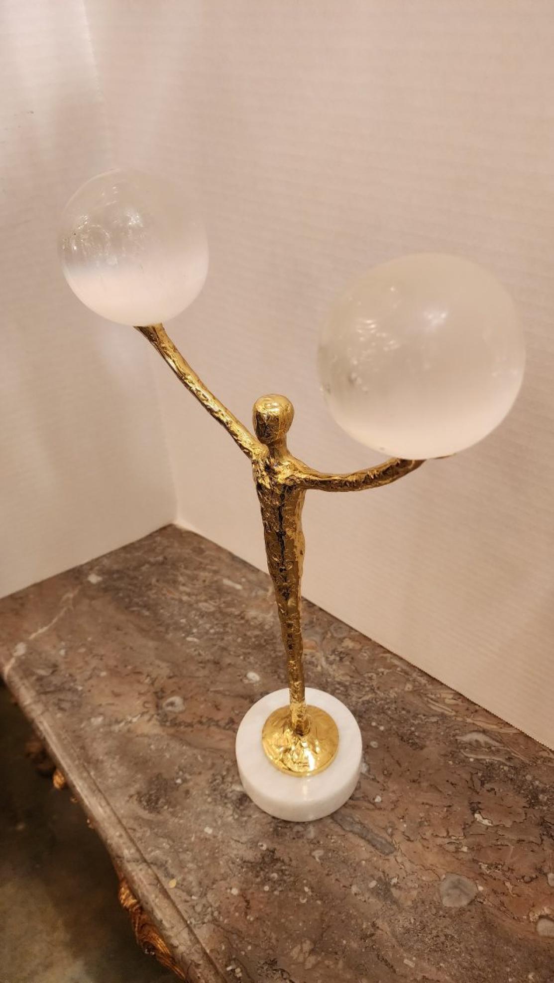 Questa inserzione presenta una scultura figurata in ottone che bilancia due sfere di selenite, in cima a una base circolare in marmo. La selenite è un cristallo affascinante noto per il suo bagliore luminoso e per le sue associazioni con la purezza,