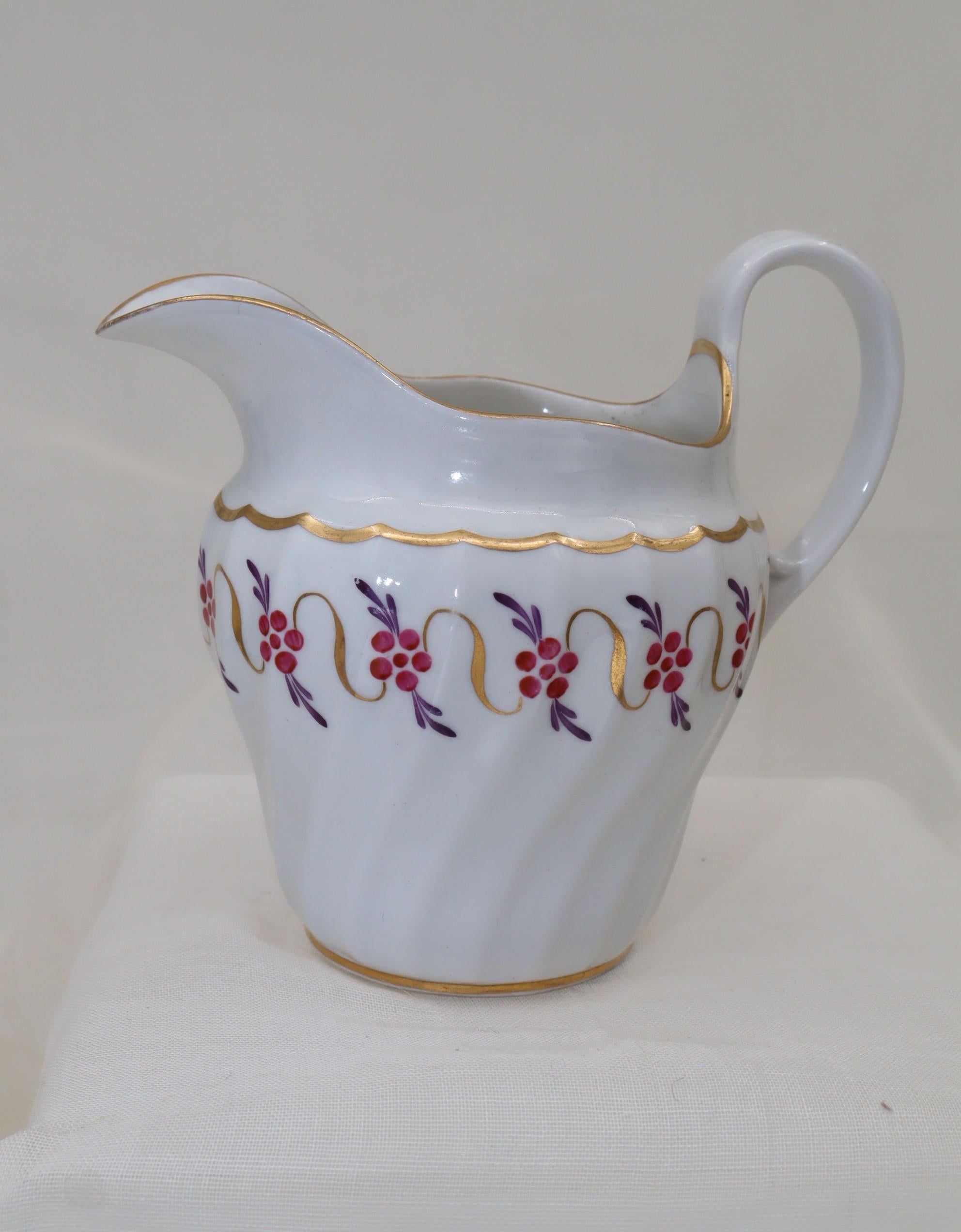 This fluted milk jug or creamer is by John Rose Coalport and is decorated with a simple repeating pattern painted in puce and purple and connected with gilt ribbons. It is further highlighted with gilding to the rim, foot, handle and the top of the
