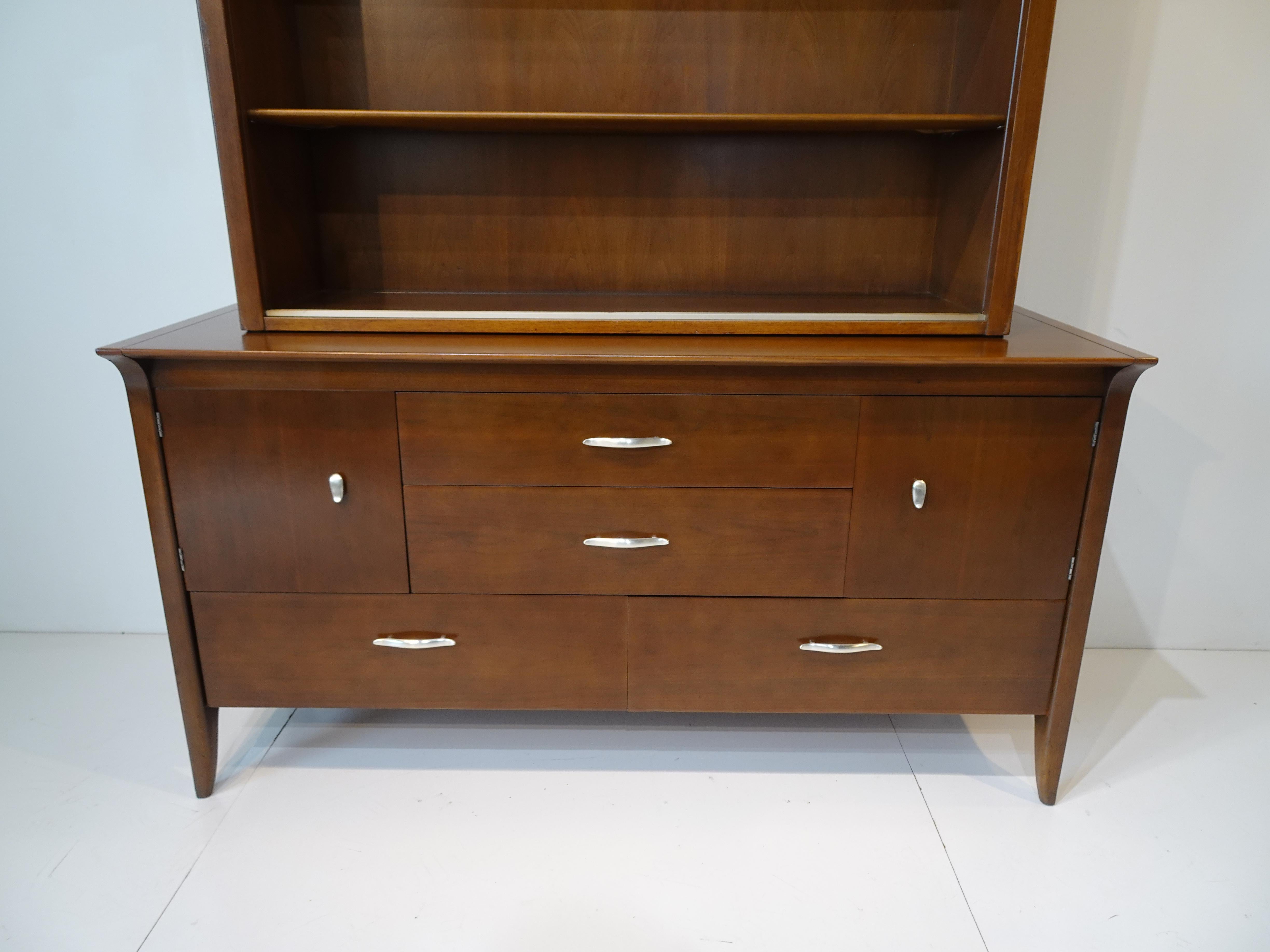 A two piece cabinet with lower sideboard having four drawers one with dividers and two doors to each side with an adjustable shelve . The sculptural pulls are in a satin chrome , the top piece has two adjustable long shelves with frosted glass