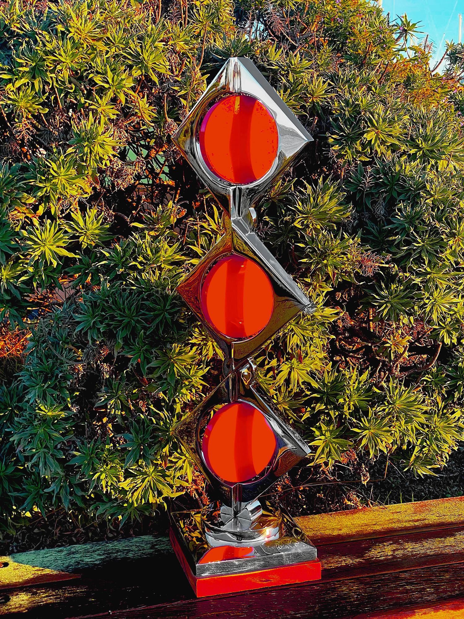 Mirror-polished stainless steel with powder-coat.

This sculpture's primary focal point is its stunning three-tier symmetric patterns. Three orange powder-coated cylindrical discs sit between three right angle, prism-shaped objects with cylindrical
