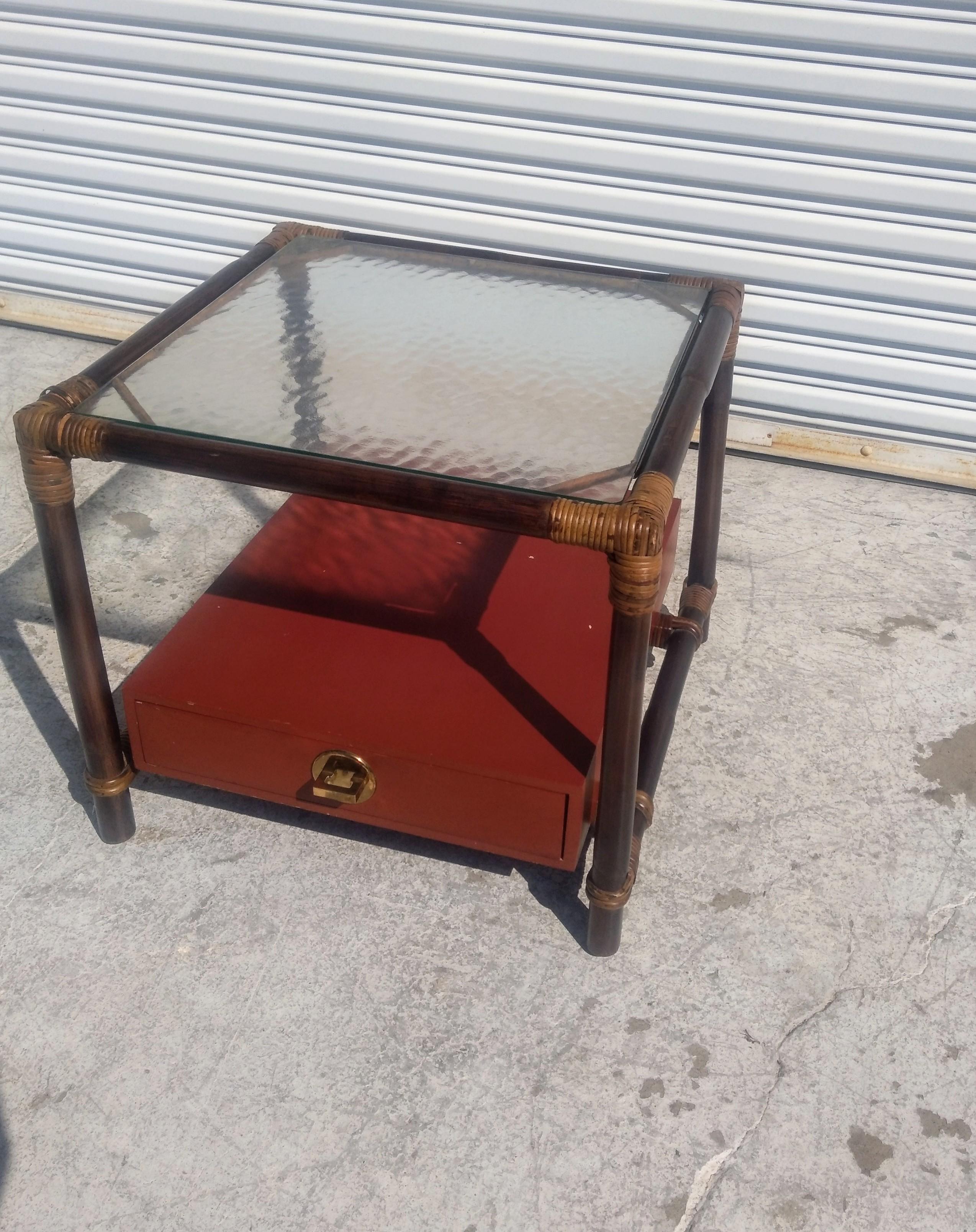 This is a nice boho chic end table by Ficks Reed. It's designed by John Wisner. It has a lower bottom drawer that also serves as a shelf for storing items. The top has wavy glass.